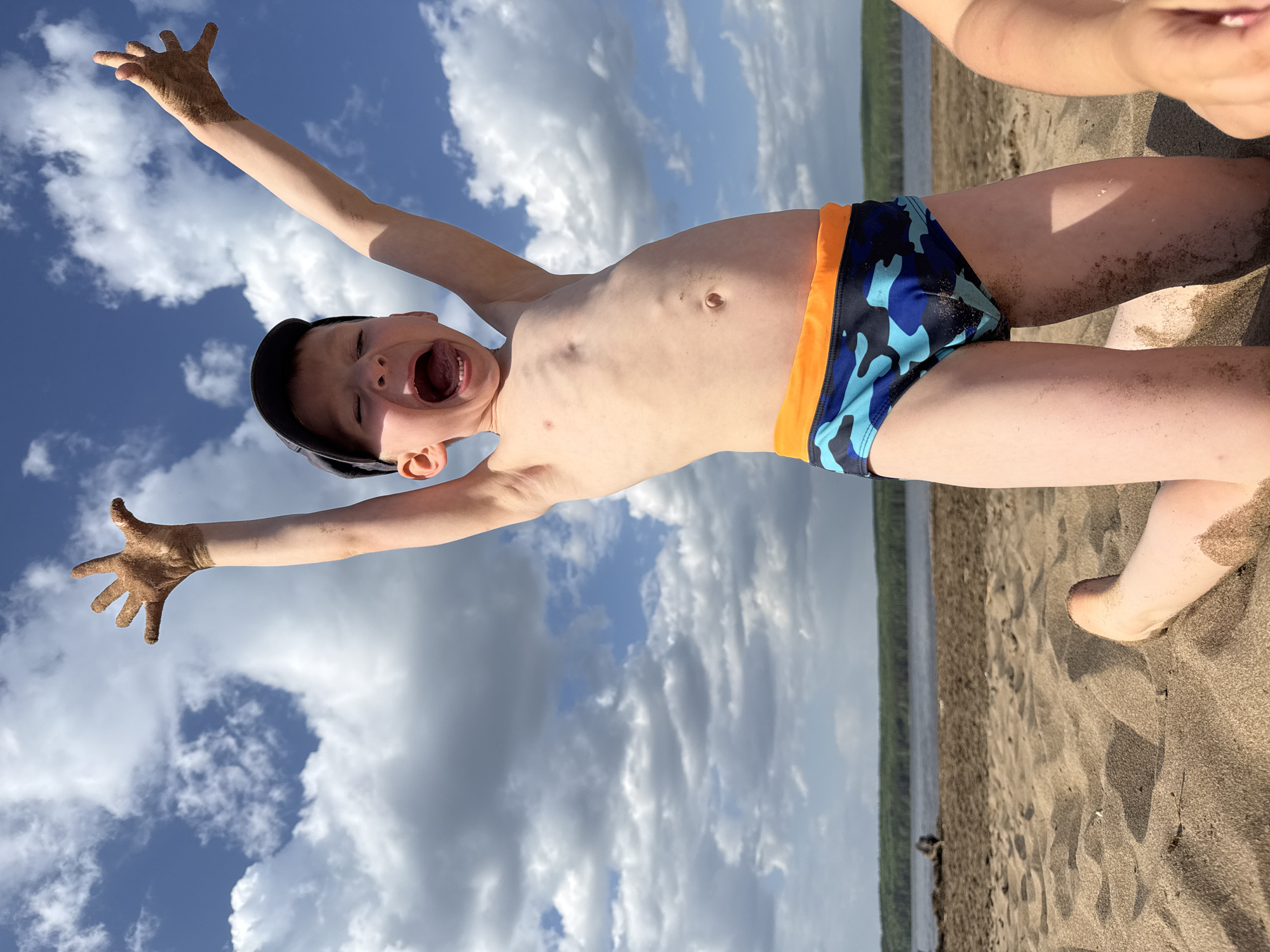 A young boy stands on a sandy beach under a cloudy sky. He wears a dark cap and blue camouflage patterned swim trunks with an orange stripe. He has his arms raised, his hands covered in sand. He is shouting with his eyes closed. The background shows water and trees. The location is Chaikovsky, Russia.