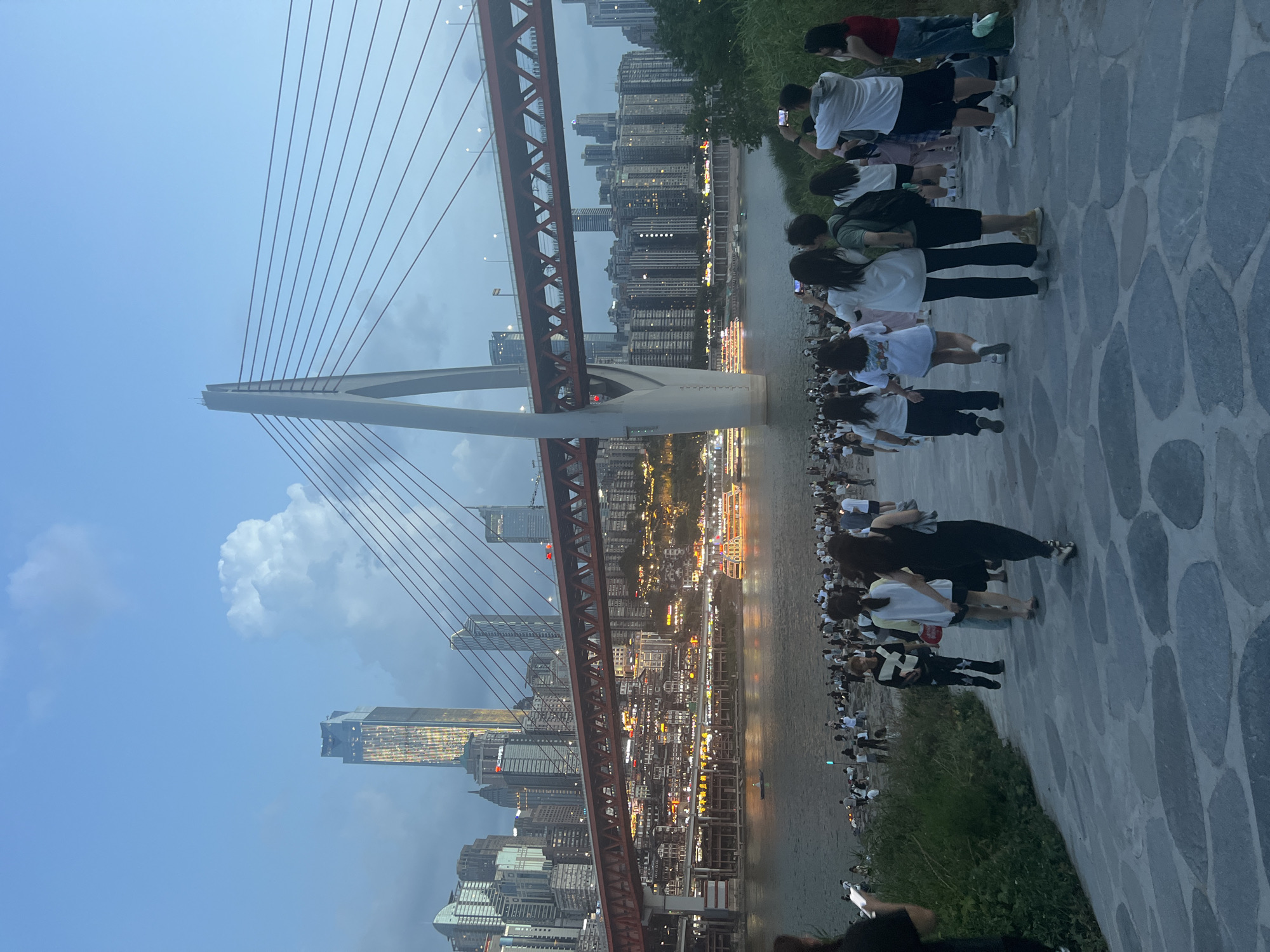 In the city of Jiangbeicheng Street, China, a crowd of people gathers along a riverbank at dusk. The scene is dominated by the striking architecture of the Yangtze River Cableway, a suspension bridge with a distinct white pylon and red suspension cables, spanning across the river. Beyond the bridge, the Chongqing skyline rises with numerous illuminated skyscrapers, creating a vibrant urban backdrop. The river itself reflects the city lights, and a few boats are visible on its surface. The foreground features a paved walkway made of irregular-shaped stones, with a grassy area to the right. People, mostly dressed in casual attire, are seen walking, standing, and taking photos with their phones, many facing the bridge and the cityscape. The sky is a dusky blue, with a large, dramatic cloud formation visible above the skyscrapers. The overall atmosphere suggests a popular evening gathering spot for locals and possibly tourists, enjoying the panoramic view of the city and its iconic bridge.