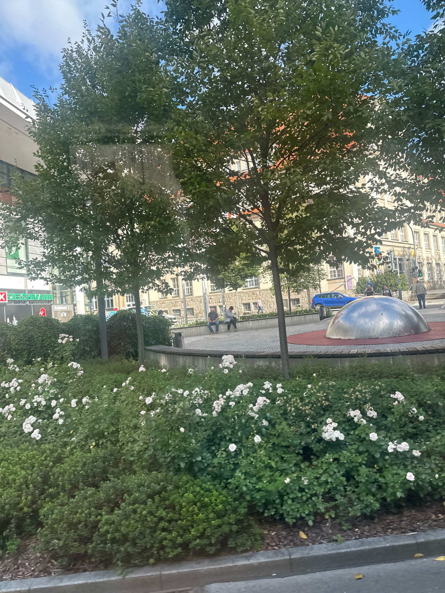 The image captures a street-level view in Bautzen, Germany, focusing on a public area with a prominent silver, dome-shaped sculpture as a centerpiece. This sculpture is situated on a circular red base within a paved square, bordered by manicured greenery and flowering white roses. Several trees with green foliage line the background, with the dappled sunlight filtering through their leaves. Behind the trees, multi-story buildings with traditional European architecture, featuring tiled roofs and numerous windows, form the urban backdrop.
In the middle ground, a few people are visible. Two individuals are seated on a concrete bench to the left of the sculpture. Further to the right, a blue car is parked, and a few pedestrians are walking. One person is sitting on a low wall to the far right. The foreground is dominated by dense, green bushes and the white rose bushes, creating a visually appealing contrast with the urban environment. A grey curb and a street are also visible at the bottom of the frame. The sky is partly cloudy, with patches of blue visible through the tree canopy. The overall impression is of a well-maintained urban public space.