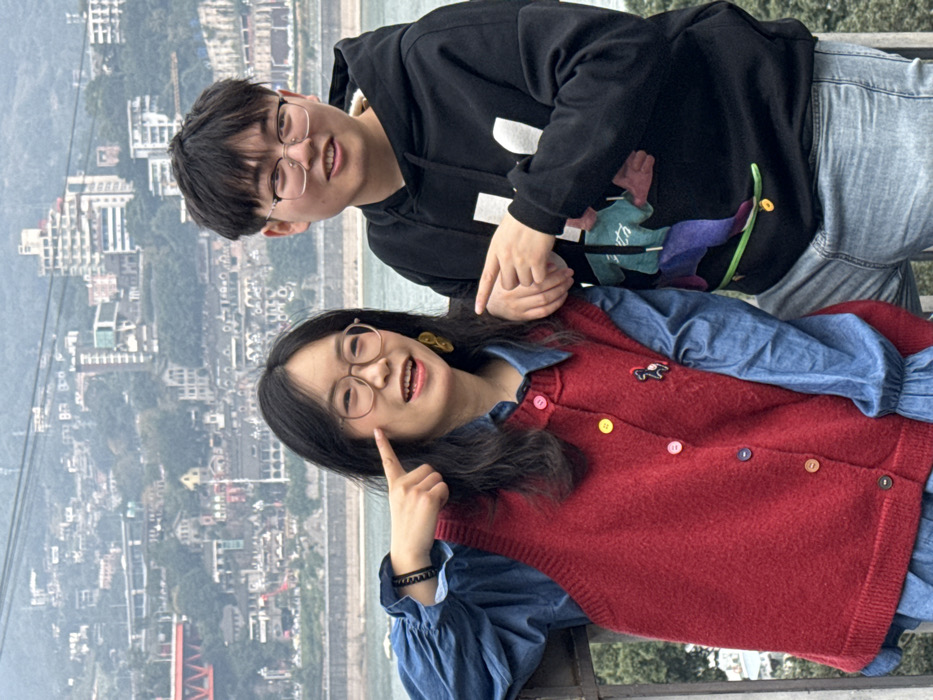 A young man wearing glasses, a black hoodie with a graphic, and jeans stands behind a young woman. The woman is pointing to her cheek and wearing glasses, a blue denim dress, and a red sweater vest with colorful buttons. They appear to be posing for a photo with a cityscape in the background. The image is rotated, with the landscape appearing sideways.