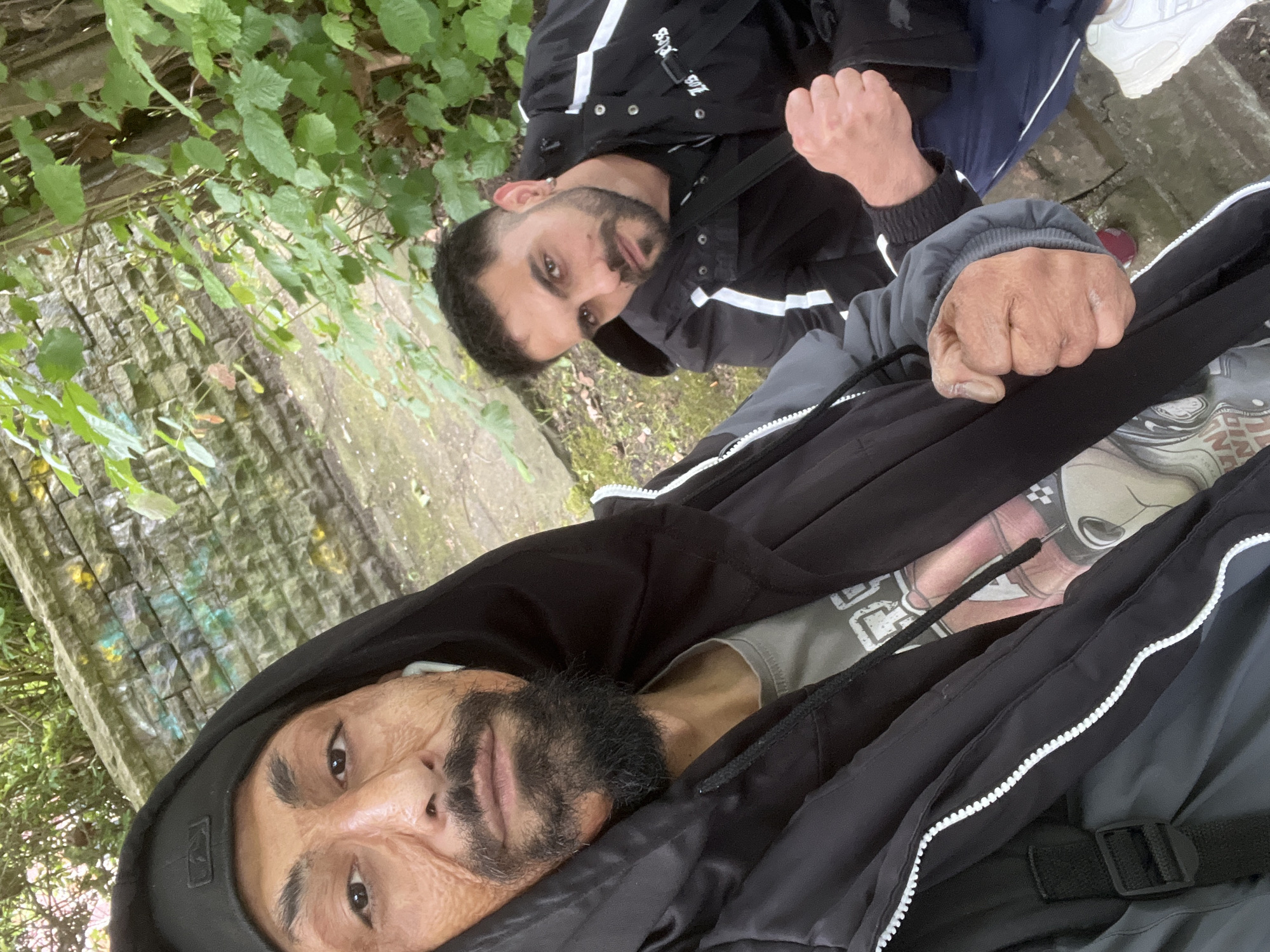 The image, captured in Herne, Germany, depicts two men in an overcast, park-like outdoor setting. The background features a weathered grey stone wall with subtle green and yellow discoloration, behind which dense green foliage is visible. A dirt path makes up the ground cover.
The man in the immediate foreground, presumed to be taking the photograph, has dark skin with prominent facial texture or scarring on his cheeks. He wears a black baseball cap, a black hooded jacket with a white zipper running down the front, and a visible grey graphic t-shirt underneath. He has a dark, pointed beard and mustache, and a white earbud in his right ear. His left hand is clenched into a fist.
The second man, positioned slightly behind and to the right, has short dark hair, a trimmed dark beard and mustache, and an earring in his left ear. He wears a black jacket with distinctive white stripes on the sleeves and front, a black t-shirt with a visible strap across his chest (suggesting a bag), dark blue track pants with a white stripe, and white sneakers. His right hand is clenched into a fist. Both individuals are looking directly at the camera with serious expressions.