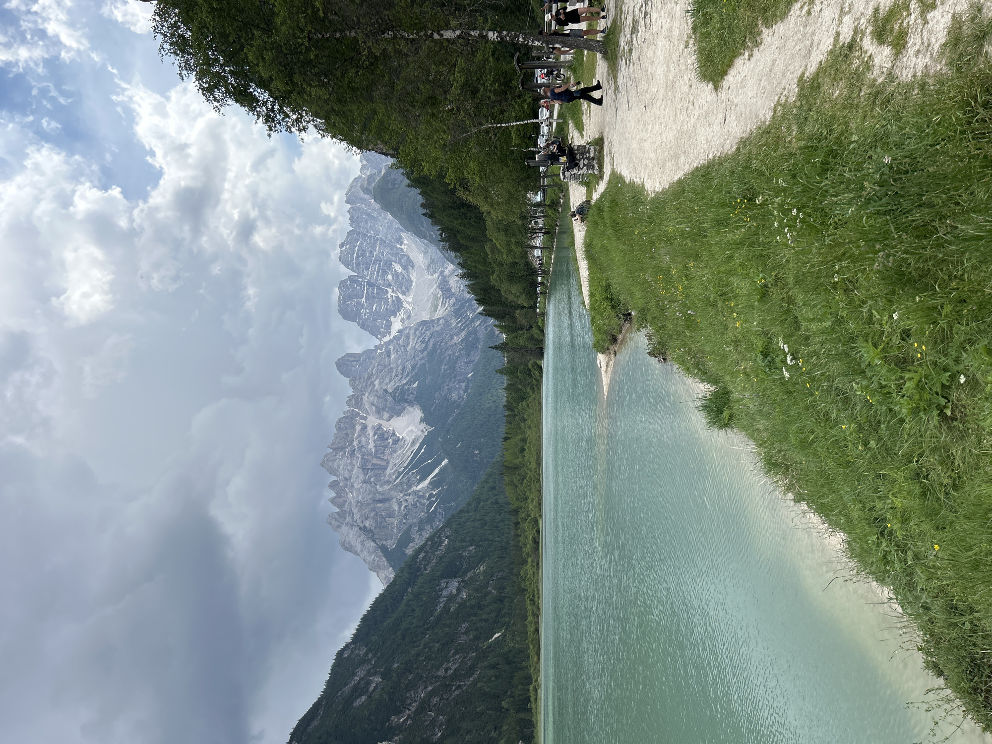 The image depicts a scenic view of a turquoise lake with snow-capped mountains in the background under a cloudy sky. In the foreground, a dirt path runs alongside the lake, bordered by lush green grass and wildflowers. Several people are visible walking and standing on the path. Trees line the edge of the path and the lake. The overall scene suggests a natural landscape, possibly a park or recreational area, with signs of human activity.