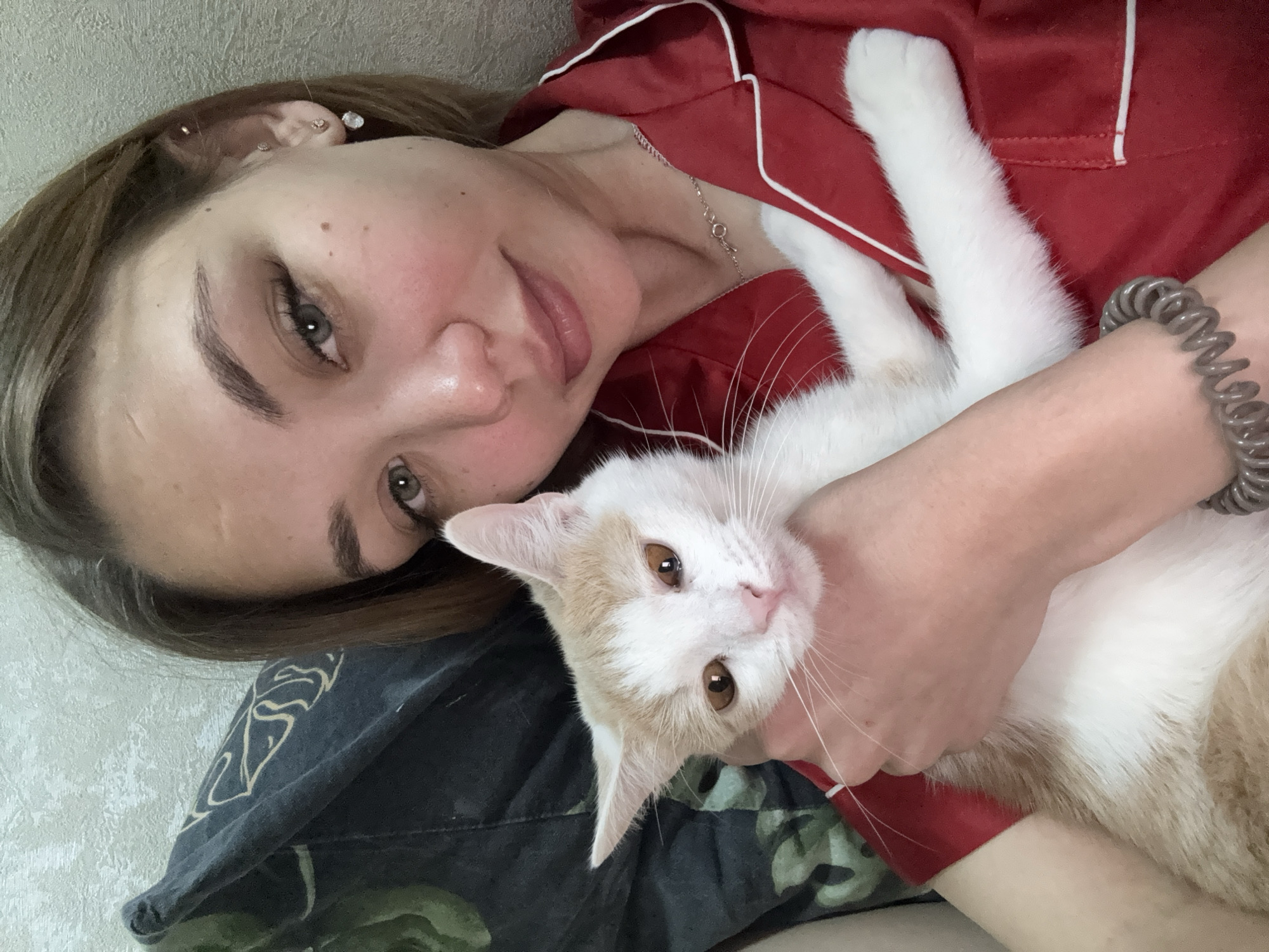 A person is lying down and holding a cat. The person has light skin, light brown hair, and green eyes. They are wearing a red pajama top with white trim and a silver necklace. The cat is mostly white with some light orange markings on its head and body. It has amber eyes and long white whiskers. The person's left wrist has a grey coiled hair tie. The background includes a pillow with a dark-colored pattern and a light-colored wall. There is no indication of this photo being taken in Kazan, Russia.