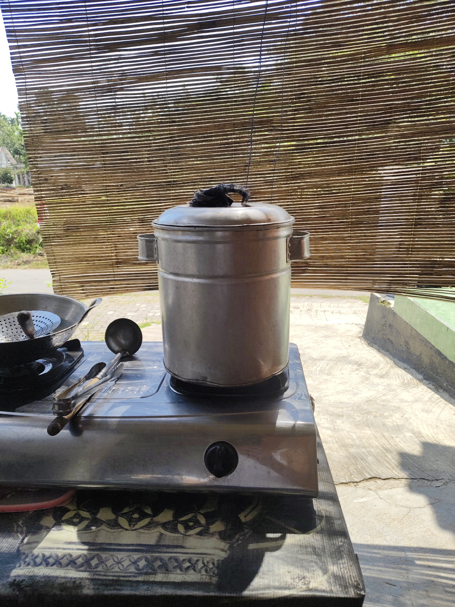 A silver pot is centered on a stainless steel gas stove, with a black ladle and tongs resting on the stove in front of it. To the left, a black frying pan with a metal strainer inside sits on another burner. The stove is placed on a dark wooden table with a decorative pattern. Behind the stove, a bamboo blind filters sunlight, revealing a blurry outdoor scene with greenery and a building in the distance. The ground appears to be concrete. The image is taken at an outdoor cooking area, possibly in Bangsalsari, Indonesia, given the context.