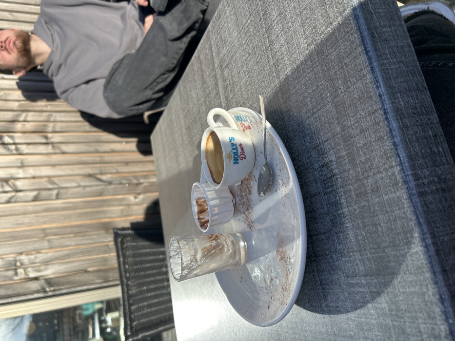 The image captures an outdoor scene, likely a patio or deck, on a bright, sunny day. The perspective is from slightly above, looking down at a table and a person. The image is rotated 90 degrees counter-clockwise, making the table appear vertical and the person horizontal.
In the foreground, a dark gray, textured rectangular table surface dominates the right side of the frame. Resting on this table is a white, oval-shaped platter. On the platter, there is a small white coffee cup filled with a light brown liquid, presumably coffee or espresso, topped with crema. The cup features blue and red lettering that reads "HOLLY'S DINER". A small silver spoon rests beside the cup on the platter. Also on the platter are a small, empty, white fluted paper cup with brown residue inside, and a clear, textured, empty glass. The platter itself has scattered brown powder, likely cocoa or coffee grounds, suggesting recent consumption.
In the background, to the left of the table, a person is visible from the chest up, seemingly leaning back or resting. The person has short dark hair and a beard or stubble and is wearing a gray hooded sweatshirt and dark jeans. Their eyes appear to be closed or looking upwards, conveying a relaxed posture. Behind the person and the table, a wooden deck or floor with horizontal planks is visible, illuminated by strong sunlight, which also casts distinct shadows across the table and on the person. Part of a dark, woven outdoor chair can be seen in the lower left portion of the image, and a faint reflection or view of greenery is visible in the very bottom left corner, possibly through a window or glass door. The strong shadows and bright light indicate it is daytime, likely morning or afternoon, with clear weather. The overall impression is a casual outdoor setting, possibly a cafe or home patio, where someone is taking a break and enjoying a drink.
