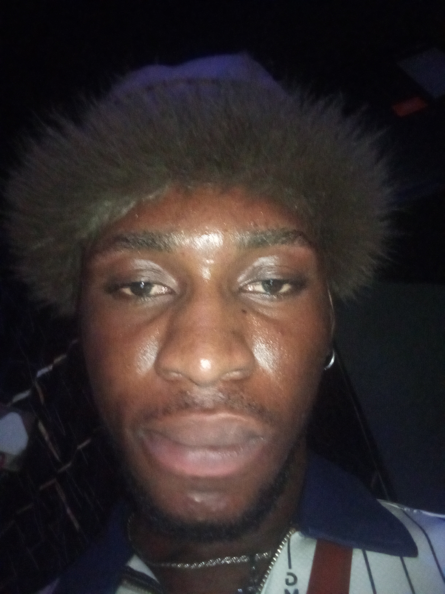 A medium shot of a dark-skinned man wearing a fur hat. He has short black facial hair and a silver hoop earring in his left ear. He is wearing a shirt with a blue collar and white and blue vertical stripes with a red stripe down the center. A silver necklace is visible around his neck. The background is dark and indistinct. The location is unknown but alleged to be Lokoja, Nigeria.