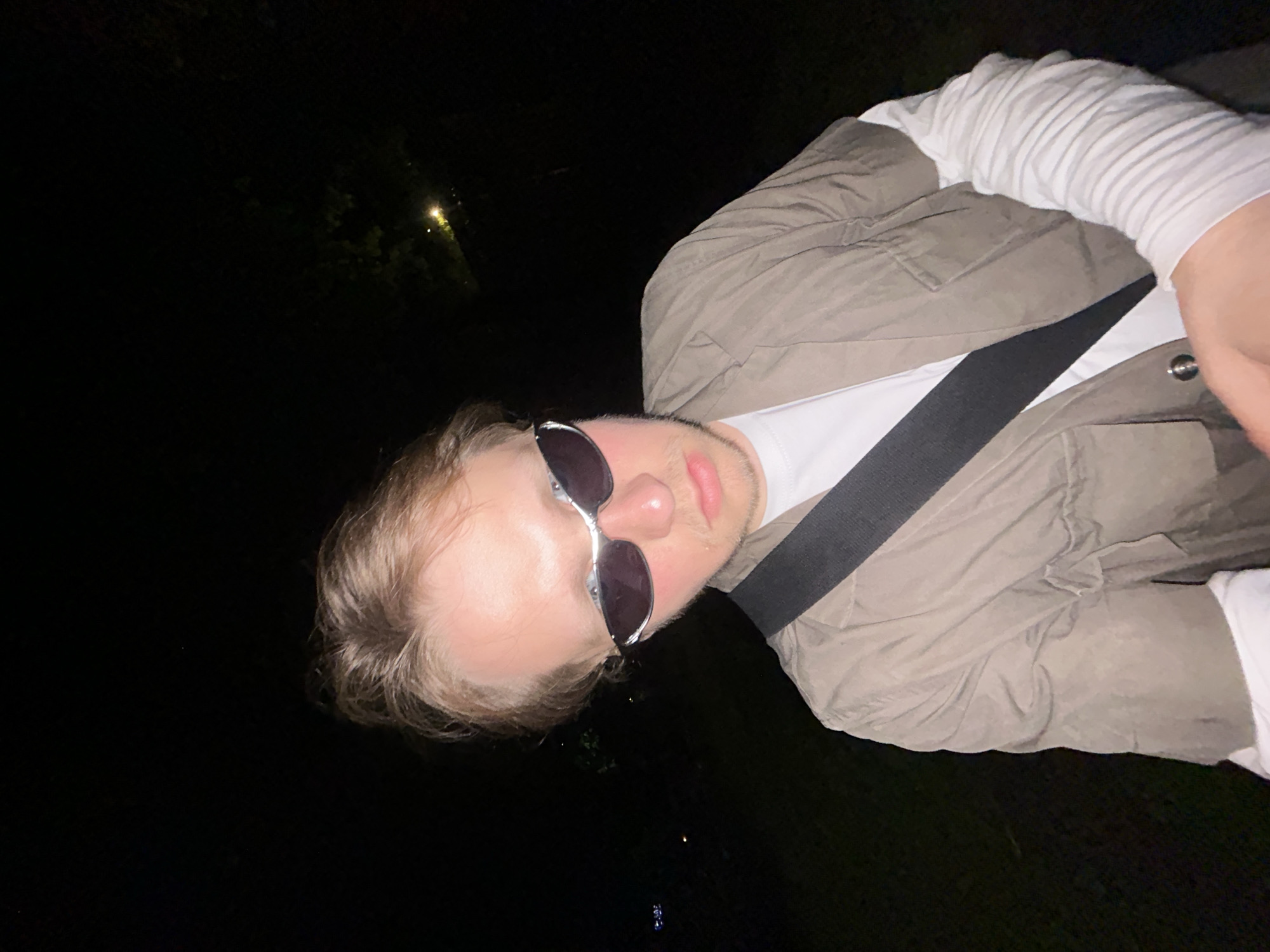 A low-angle, canted, medium shot captures a person wearing sunglasses, a white t-shirt, a beige jacket, and a grey ribbed long-sleeved undershirt. A grey strap bisects the frame diagonally from the upper-left to the lower-right, crossing over the person's chest. The background is predominantly black, indicating a nighttime setting, with indistinct illumination suggesting a city environment in the distance. The location and specific city are not discernible.
