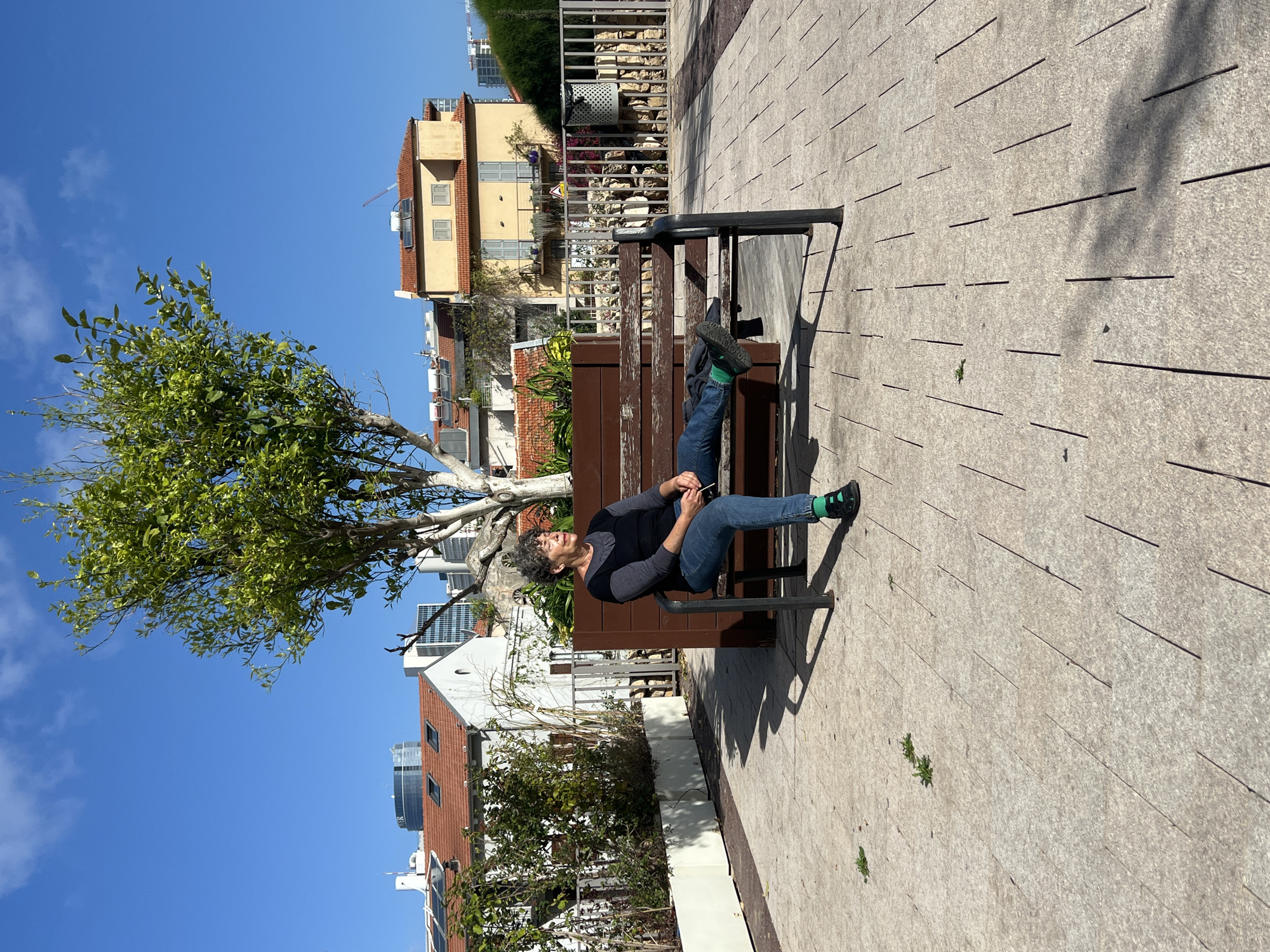 A woman with short gray hair is reclining on a brown wooden bench in what appears to be an urban park or walkway in Tel Aviv, Israel. She is wearing a black top, blue jeans, and dark shoes with bright green socks. Her legs are extended, with her feet resting on the far end of the bench. She appears to be holding something in her hands.
Behind the bench, the scene includes greenery, trees, and a variety of buildings, including residential homes with red-tiled roofs and modern structures. The sky is a clear blue with a few scattered clouds. The ground is paved with textured, light-colored concrete or stone tiles. The photo is taken during daylight hours, casting shadows from the bench and the tree to the left.