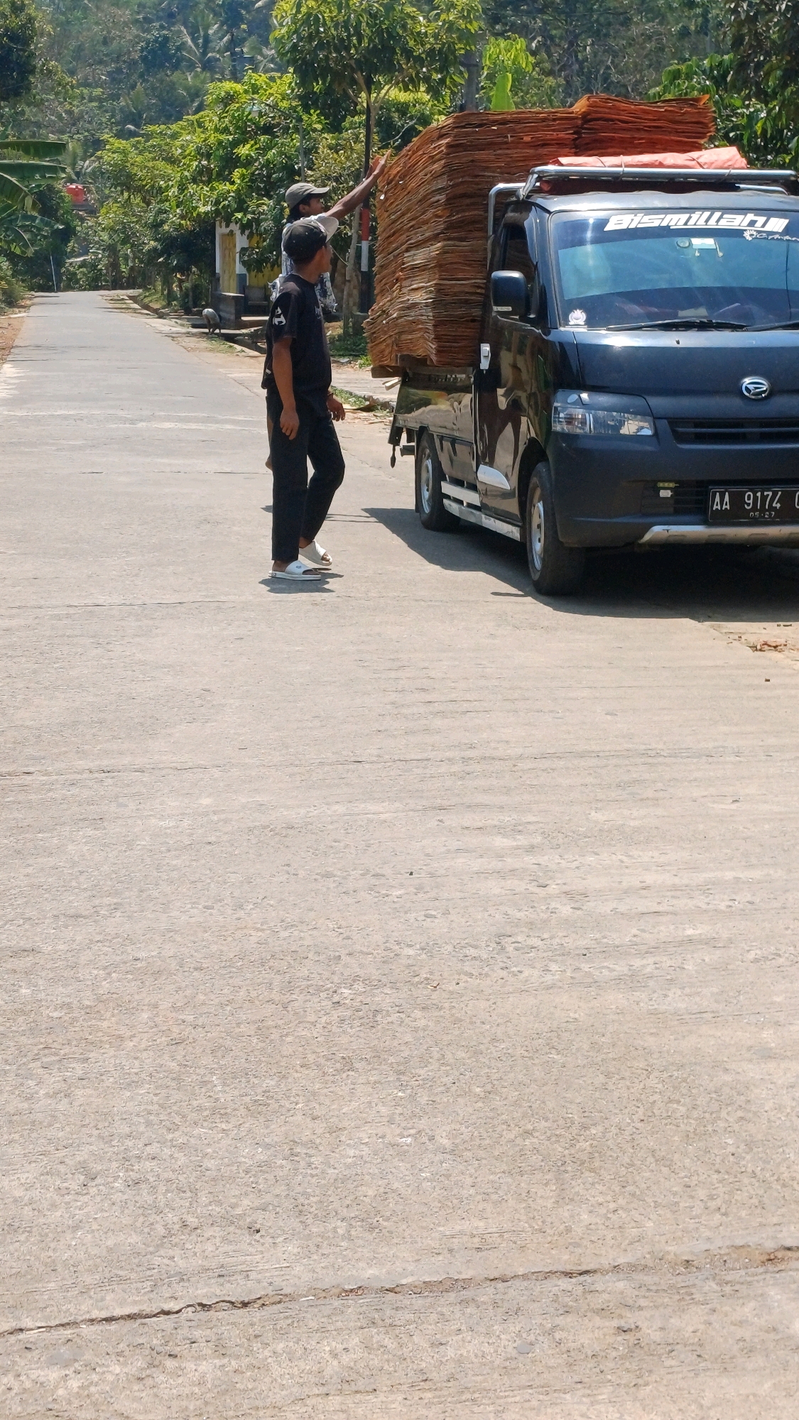 In the city of Pringsurat, Indonesia, on a paved road with some cracks, two men are seen near a black pickup truck loaded with stacks of what appears to be corrugated cardboard or wood planks. The truck has a silver bumper and a white sticker on the windshield with the word "Bismillah". One man, wearing a black t-shirt and black pants with white slides, stands facing the truck. The other man, wearing a white t-shirt and a dark cap, stands behind the first man, reaching towards the top of the load on the truck. The road is lined with greenery and trees, suggesting a rural or semi-rural setting. The sky is bright and clear, indicating a sunny day. The license plate of the truck is visible, reading "AA 9174".