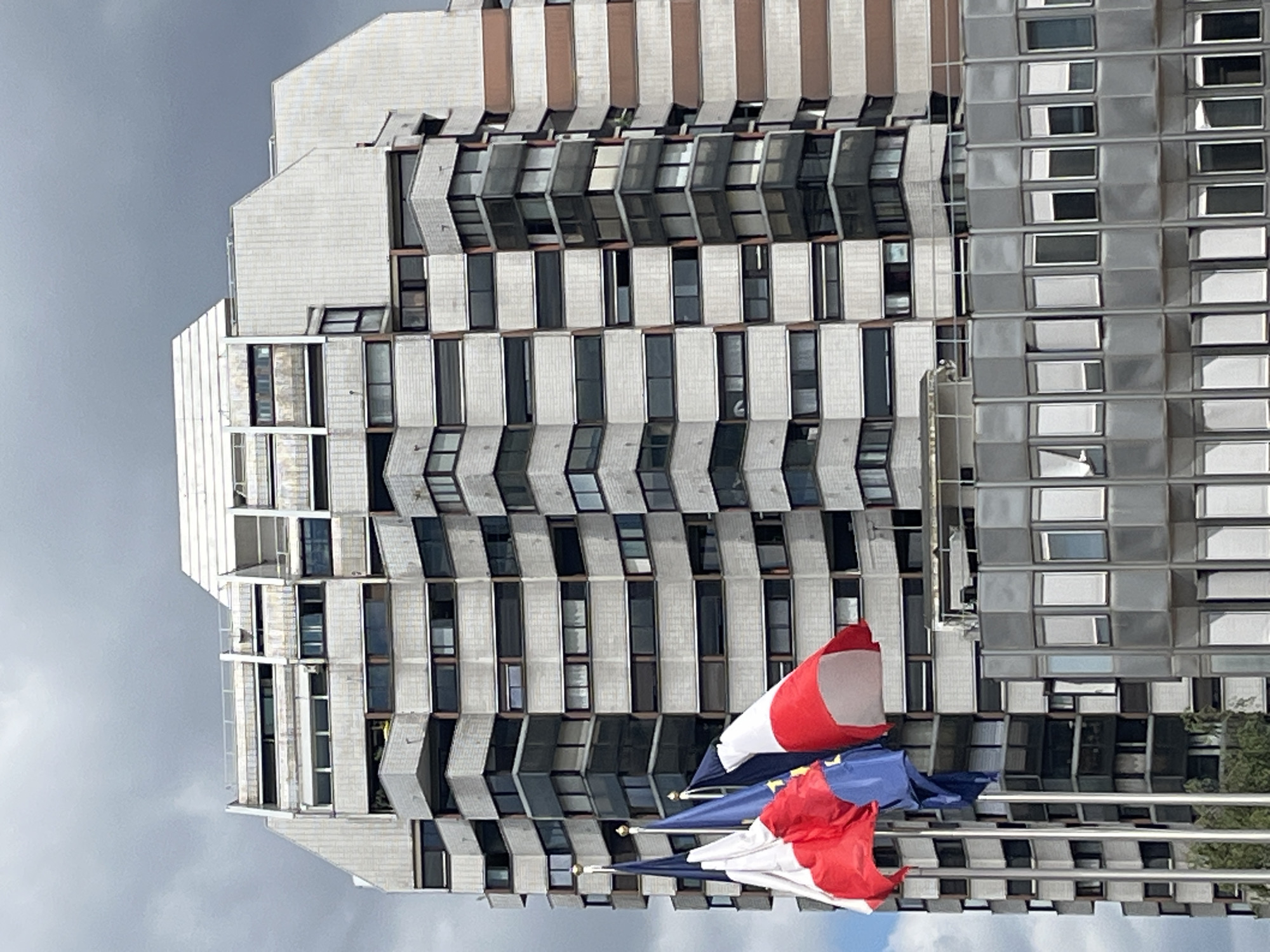 The image shows a close-up of the exterior of a modern building, likely an apartment complex or office building, in Créteil, France. The building is tall and has a distinctive architectural style with numerous balconies and large windows. The facade appears to be constructed from a combination of grey concrete or stone panels and dark-tinted glass. There are horizontal bands of a lighter, possibly beige or brown, material running vertically up the right side of the building.
In the foreground, three flags are prominently displayed on flagpoles. Two of the flags are the French tricolor (blue, white, and red), and the third flag is blue with yellow stars, representing the European Union. The flags are partially obscuring the lower section of the building. The sky above is overcast with grey clouds, suggesting a cloudy or potentially rainy day. The overall impression is of a contemporary urban environment.