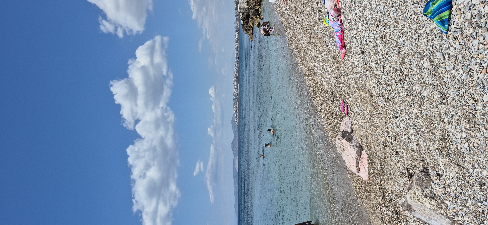 The image captures a sunny day at a pebble beach in Paleo Faliro, Greece. The sea is calm and clear, with several people swimming and wading in the shallow water. In the background, a cityscape is visible along the coastline, with buildings extending across the horizon. White, fluffy clouds are scattered across a bright blue sky. On the beach, a few towels and flip-flops are laid out, suggesting recreational activity. The shoreline is covered in small pebbles. The overall scene depicts a typical summer day at the beach.