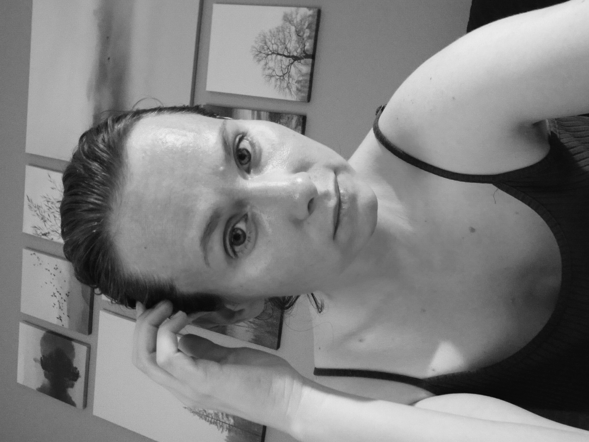 A black and white, medium shot photograph captures a woman with dark hair pulled back, her left hand near her temple as she looks directly at the camera. Her face is slightly flushed and shiny, possibly from sweat or product. She wears a dark, thin-strapped top. In the background, several landscape artworks are mounted on a gray wall. The lighting suggests an indoor setting. The image does not provide any specific geographic markers to indicate a location in Yekaterinburg, Russia.