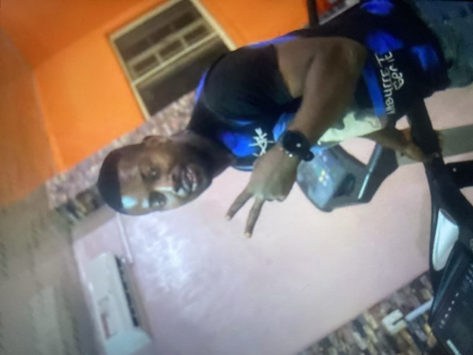 A man poses indoors, making a peace sign with his right hand. He wears a black t-shirt with blue accents and a wristwatch. Behind him, a window with a metal grid is visible against an orange wall. Below him, a white air conditioning unit is mounted on a pinkish wall. An exercise machine is to his right. The image is slightly out of focus and appears rotated.