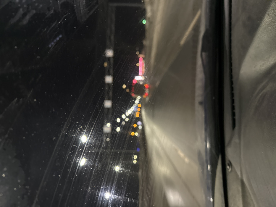 The image presents a nighttime scene captured from inside a vehicle on a multi-lane highway in Kukushtan, Russia. The view through the windshield is partially obscured by accumulated dirt, streaks, and particulate matter. The vehicle's dashboard and the base of the windshield wipers are visible in the lower portion of the frame. The road ahead is wet and highly reflective, illuminated by numerous white streetlights that appear as blurred circular light sources. Multiple vehicles are present on the highway, indicated by their distant, blurred red taillights and yellow/orange headlights. A distinct set of red taillights suggests a larger vehicle, possibly a truck or bus, in the mid-ground. Overhead gantry structures, potentially holding signs or cameras, are visible further down the road. The immediate roadside, particularly on the right, shows evidence of snow accumulation. No people are visible within the frame.
