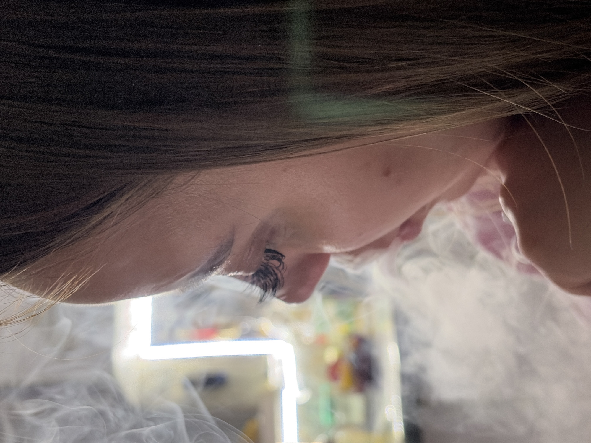 The image shows a close-up of a person's face, specifically the eye, nose, and mouth area, from an upside-down perspective. The person has dark hair and long eyelashes. The lower part of the face is obscured by what appears to be smoke or vapor. In the background, there is a blurred, illuminated rectangular object, possibly a light fixture, and some other indistinct shapes with colors. There's nothing that indicates it's in Port-Katon, Russia.