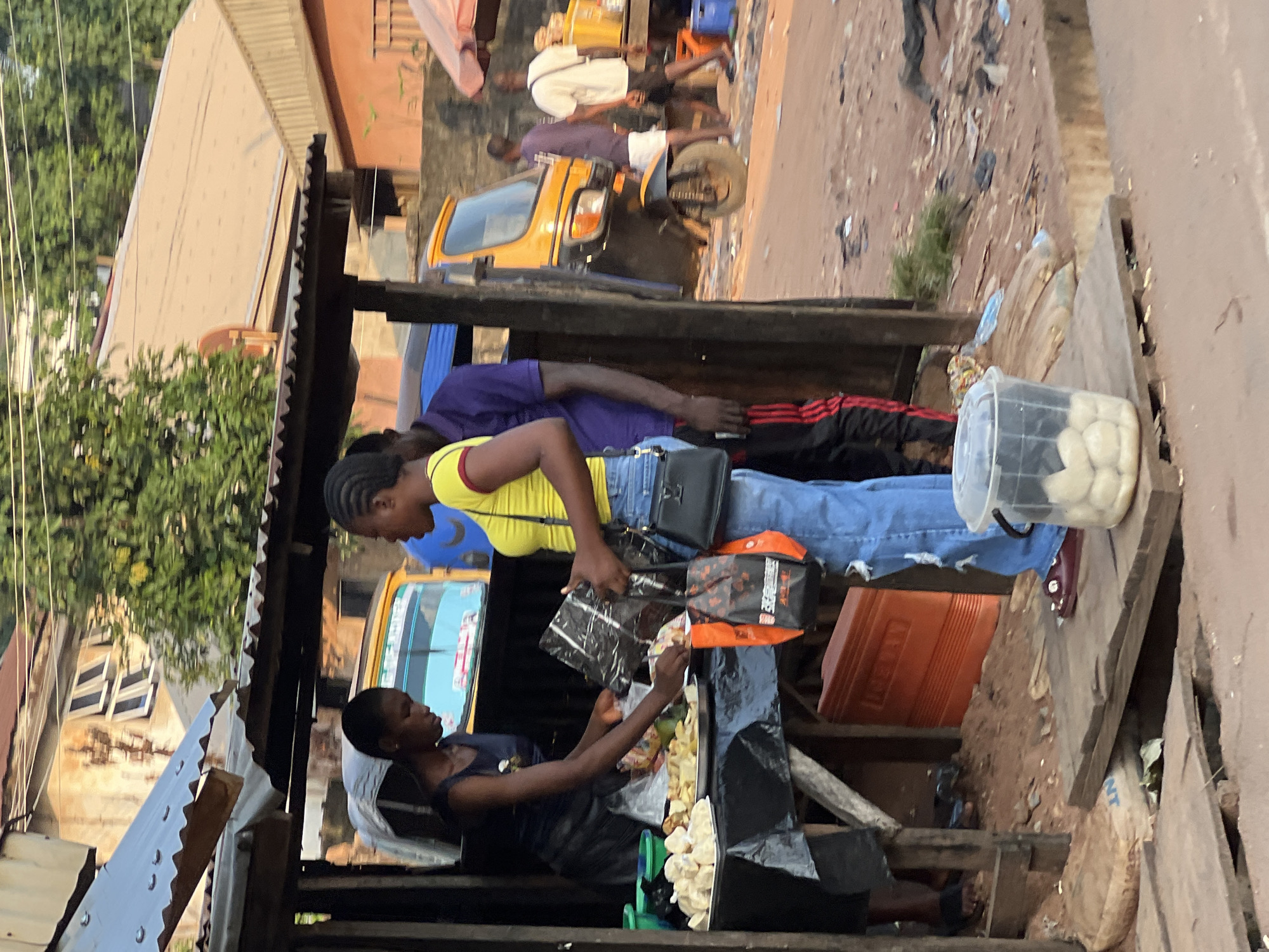 This is a medium shot capturing a street scene in Nkpor, Nigeria. In the foreground, a young woman in a yellow top and blue jeans stands by a makeshift stall. She is holding a black plastic bag and looking down at a table laden with food items. To her left, another young woman in a dark blue sleeveless top and black skirt is serving food. Behind them, a yellow three-wheeled vehicle, commonly known as a "Keke," is visible. The stall appears to be constructed from wood and corrugated metal. The ground is unpaved and littered with debris. In the background, buildings with corrugated metal roofs and a tree are visible. The lighting suggests it is daytime.