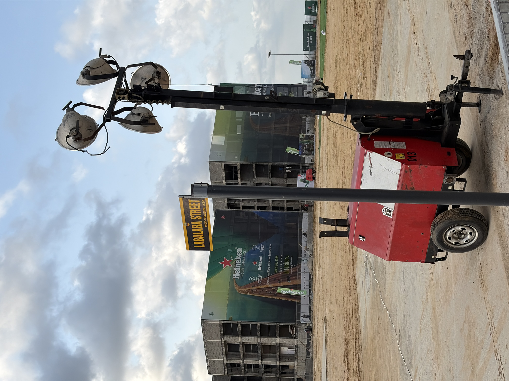 The image depicts a construction site in Lagos, Nigeria, as indicated by the "LABALABA STREET IKOYI-OBALENDE LCDA" sign. There is a portable light tower with four lamps, painted in red and white. Behind the light tower, a large billboard promoting Heineken beer and the UEFA Champions League is displayed on the facade of a partially constructed building. The billboard advertises "Unmatched View Exclusive Experiences" from May 3-9, 2026. The foreground consists of a dusty or sandy surface. The sky above shows a mix of blue and cloudy patches. In the background, there are more construction sites and advertisements.