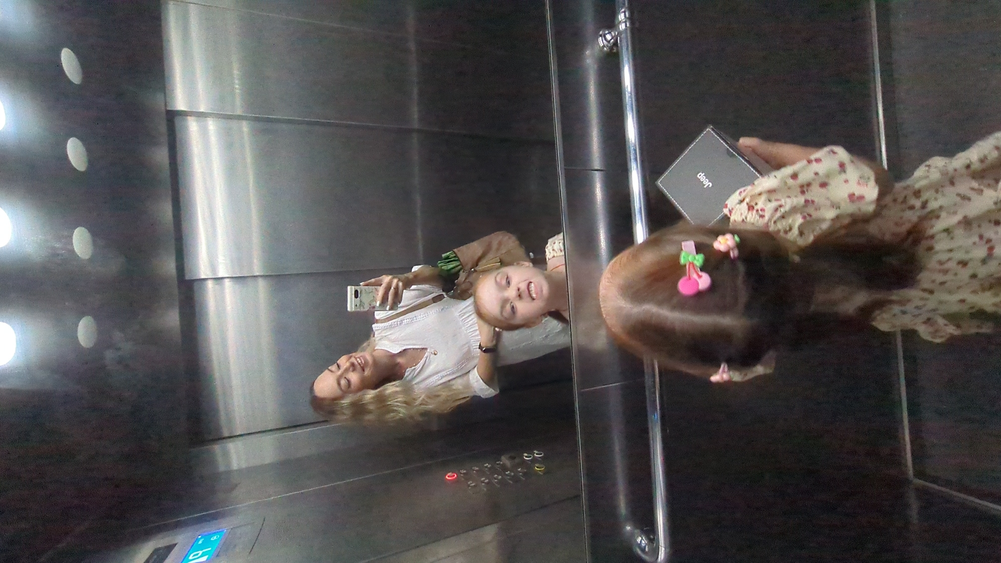 In an elevator with brushed metal walls and recessed ceiling lights, a woman with long blonde hair takes a selfie in the mirror. She is smiling and wearing a white dress with a V-neck. In the reflection, a young girl with red hair and colorful hair clips is making a face at the camera. The girl is wearing a light-colored dress with a floral pattern. In her hands, she holds a small black box with the word "Jeep" printed on it. The elevator has a control panel with red illuminated buttons and a blue digital display. A metal handrail runs along the side of the elevator. The scene is captured from a low angle, focusing on the girl's back and the reflection of the woman and girl in the mirror.