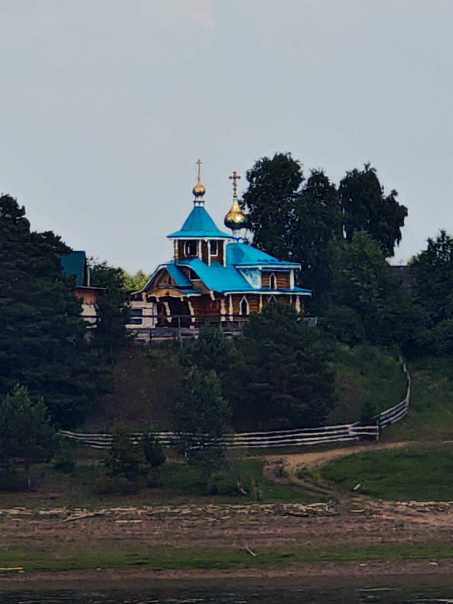 The image depicts an Orthodox church situated on a tree-covered hill overlooking a body of water. The church is constructed of wood and features a bright blue roof with golden domes, topped with crosses. A wooden fence encloses a path leading up to the church, which appears to be the only structure visible in the immediate vicinity. The surrounding environment is lush with trees and greenery, suggesting a rural or natural setting. The sky above is overcast and a light gray. The time of day appears to be either early morning or late evening due to the soft lighting. No people are visible in the image. This scene is located in Erkalovo, Russia.
