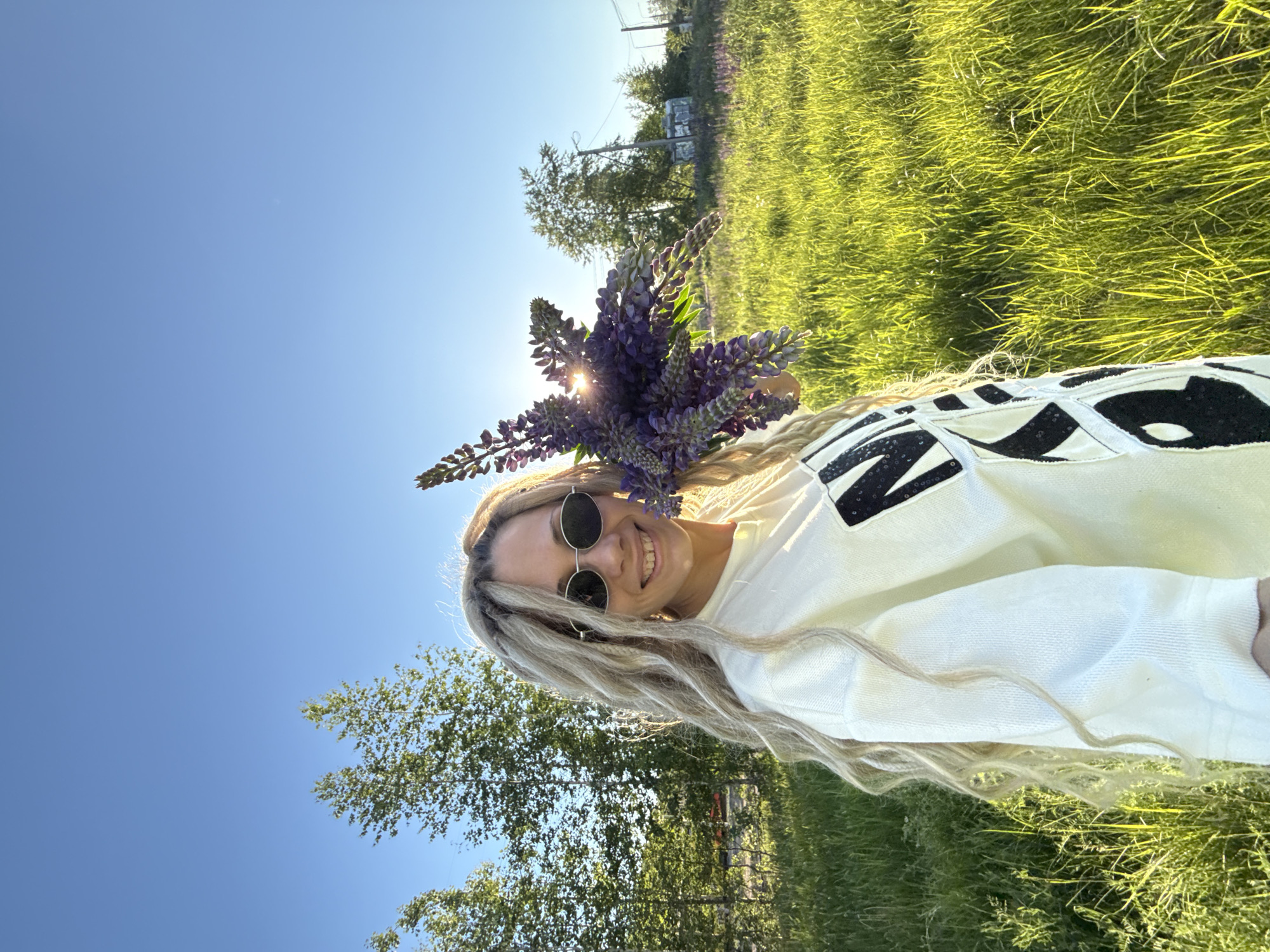 An outdoor, eye-level shot captures a female with long, blonde, wavy hair and partial dreadlocks, smiling directly at the camera. She wears dark, oval-framed sunglasses and a light-colored long-sleeved top with black patterns. Her left arm holds a bouquet of purple flowers, likely lupines, which partially obscure the sun, creating a starburst effect. The background features a vibrant green grassy field, with several trees, possibly birches, on the left and right. In the far right background, utility poles, wires, and a few distant structures are visible under a clear, bright blue sky. The scene is illuminated by strong sunlight from the right, creating highlights and backlighting. The image is set in the city of Unknown, Unknown.