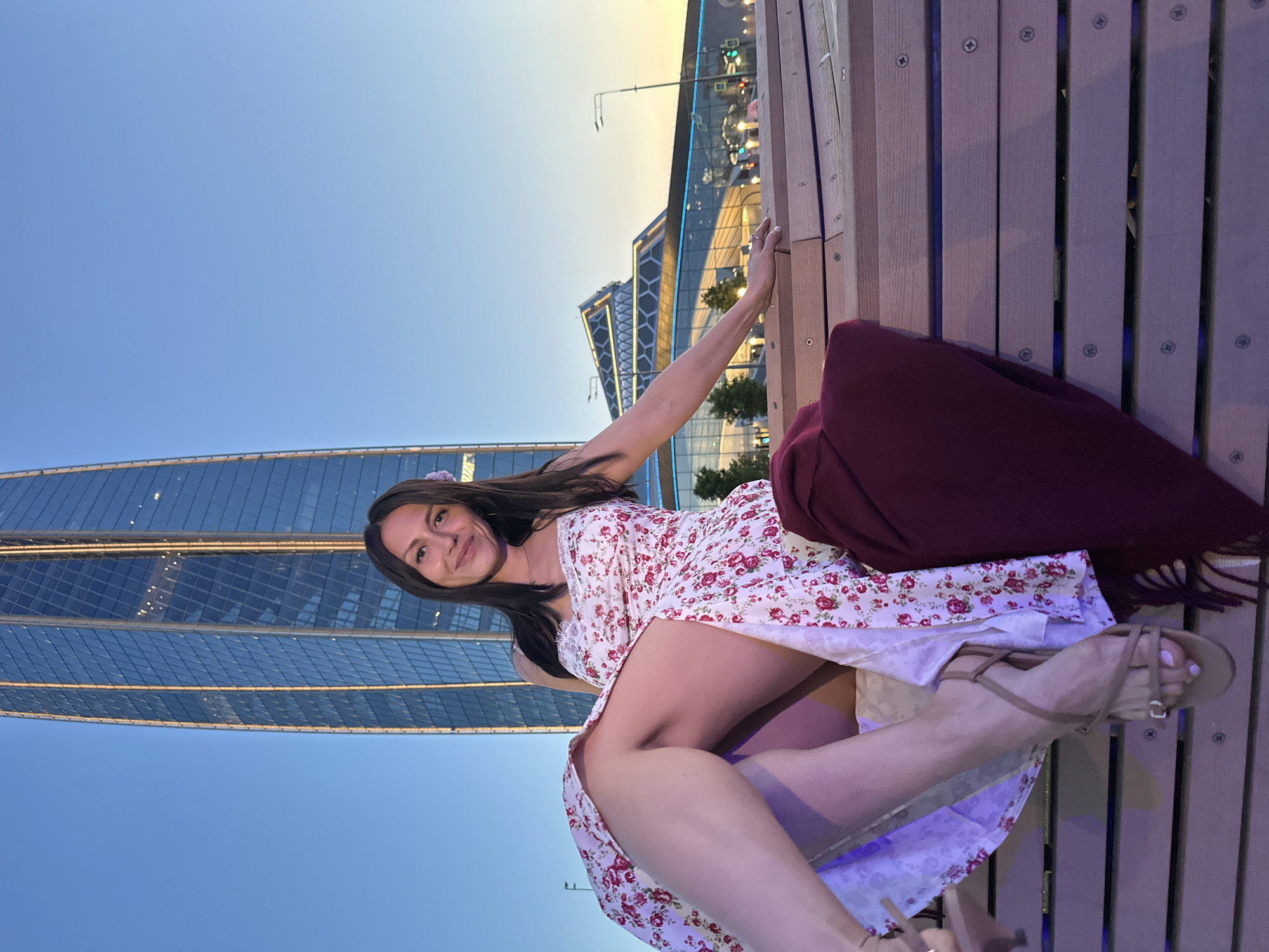 A woman is sitting on a wooden bench in front of a modern glass skyscraper at dusk. She is wearing a floral sundress and sandals, and a maroon scarf is draped over the bench beside her. The skyscraper, which appears to be Lakhta Center, is illuminated by vertical lights and reflects the twilight sky. In the background, another building with a distinctive geometric design is also lit up. The scene suggests an urban setting in Saint Petersburg, Russia, during the evening.
