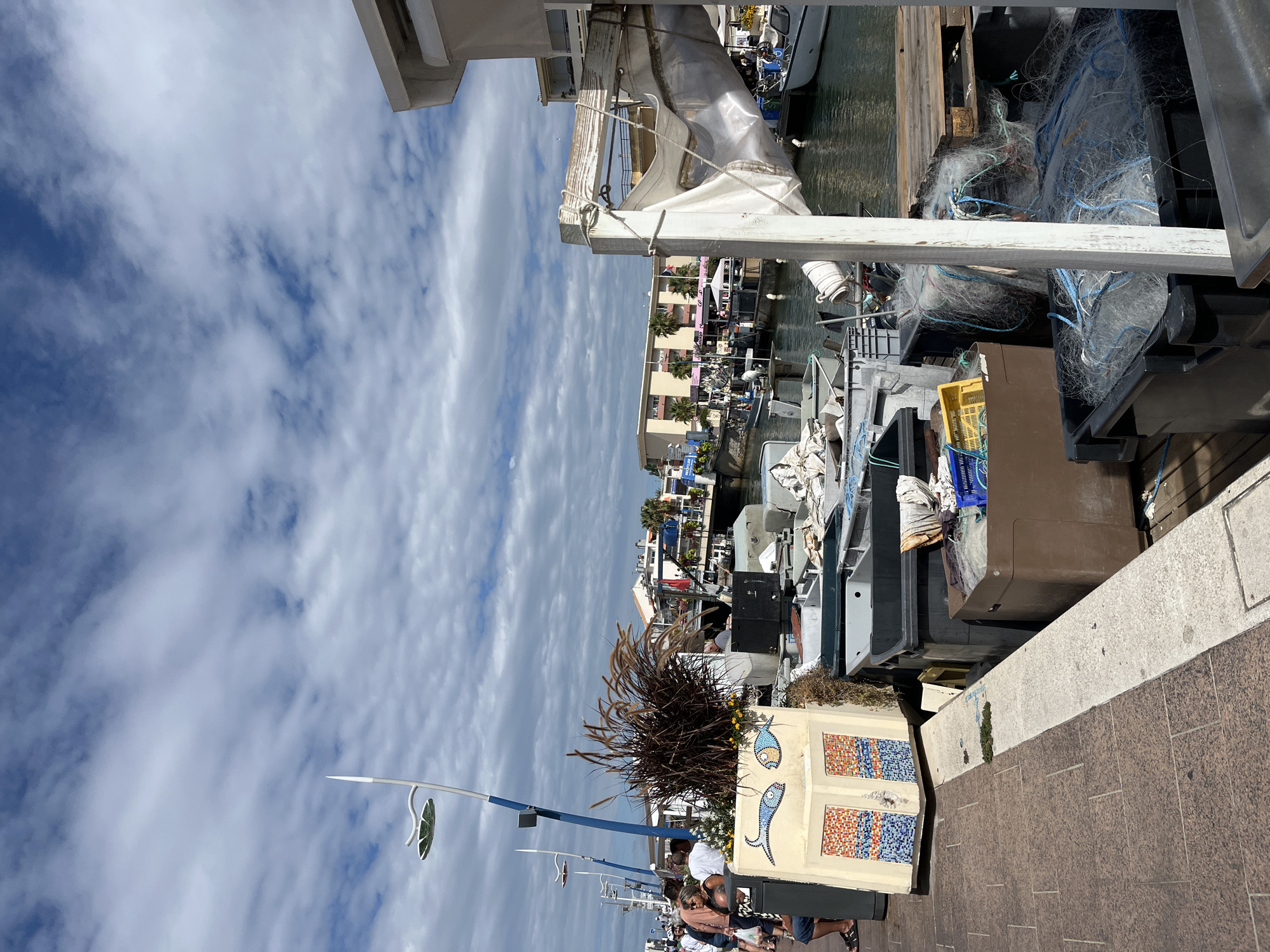 The image depicts a sunlit scene in Palavas-les-Flots, France, likely near the port, with a canal or harbor filled with boats. In the foreground, there's a paved walkway with people strolling. A decorative planter with a mosaic of fish and colorful tiles stands prominently. Adjacent to it, a collection of large grey and brown plastic bins are visible, some containing fishing nets and other equipment. A white wooden structure with some draped fabric is also in the foreground, obscuring part of the view. In the background, a row of buildings with a Mediterranean architectural style lines the waterfront, and several boats are moored in the canal. The sky is partly cloudy, with cumulus clouds against a blue backdrop. The overall impression is of a bustling coastal town on a pleasant day.