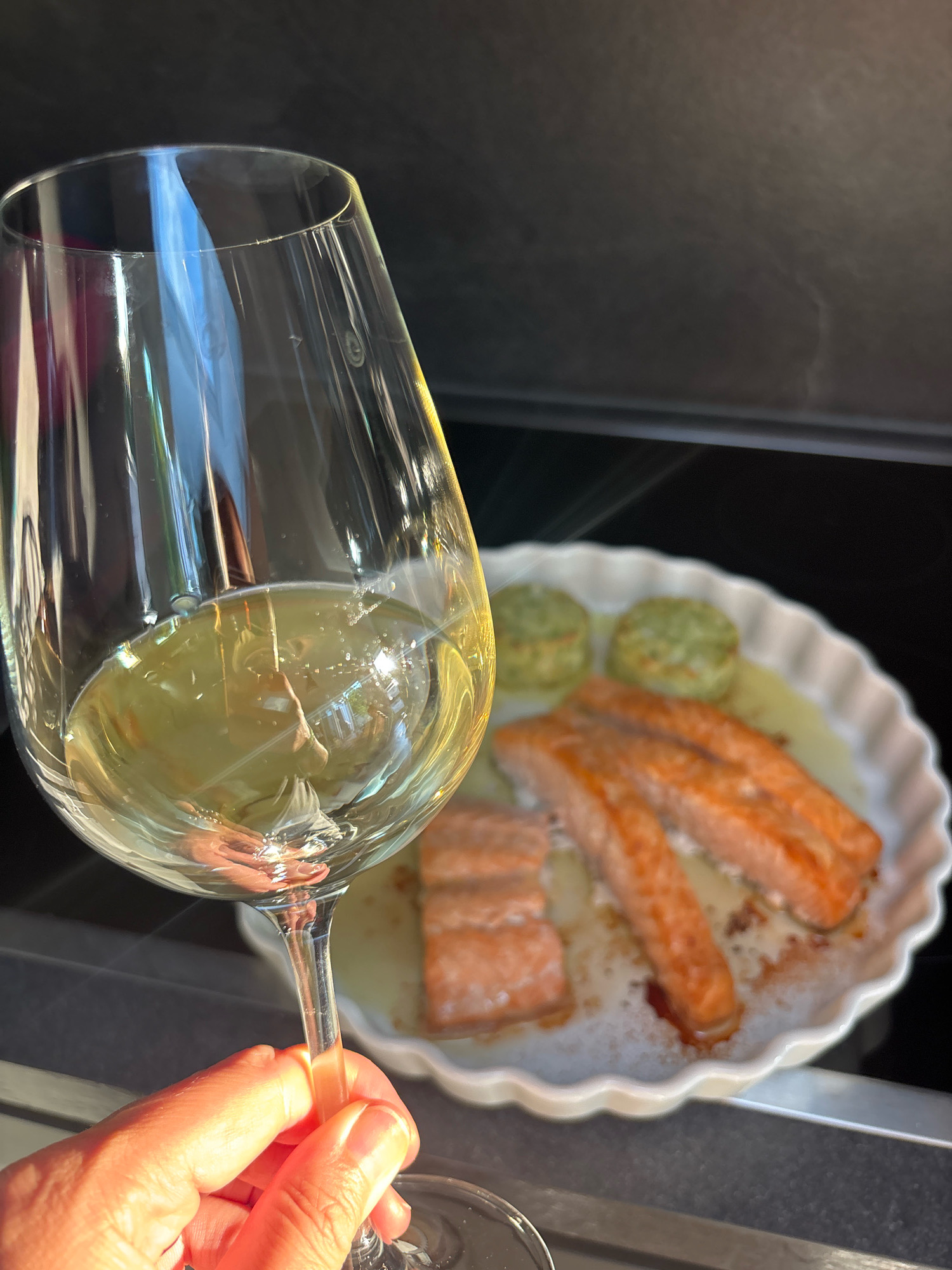 A hand holds a glass of white wine, the glass angled to show the amber liquid and reflections. In the background, a white ceramic dish contains cooked salmon fillets and two green, circular items, possibly a side dish. The scene appears to be indoors, with a stovetop visible in the lower portion of the image. The lighting suggests a sunny day, with strong highlights and shadows. The location in Frankfurt, Germany, is not discernible from the visual information present in the image.
