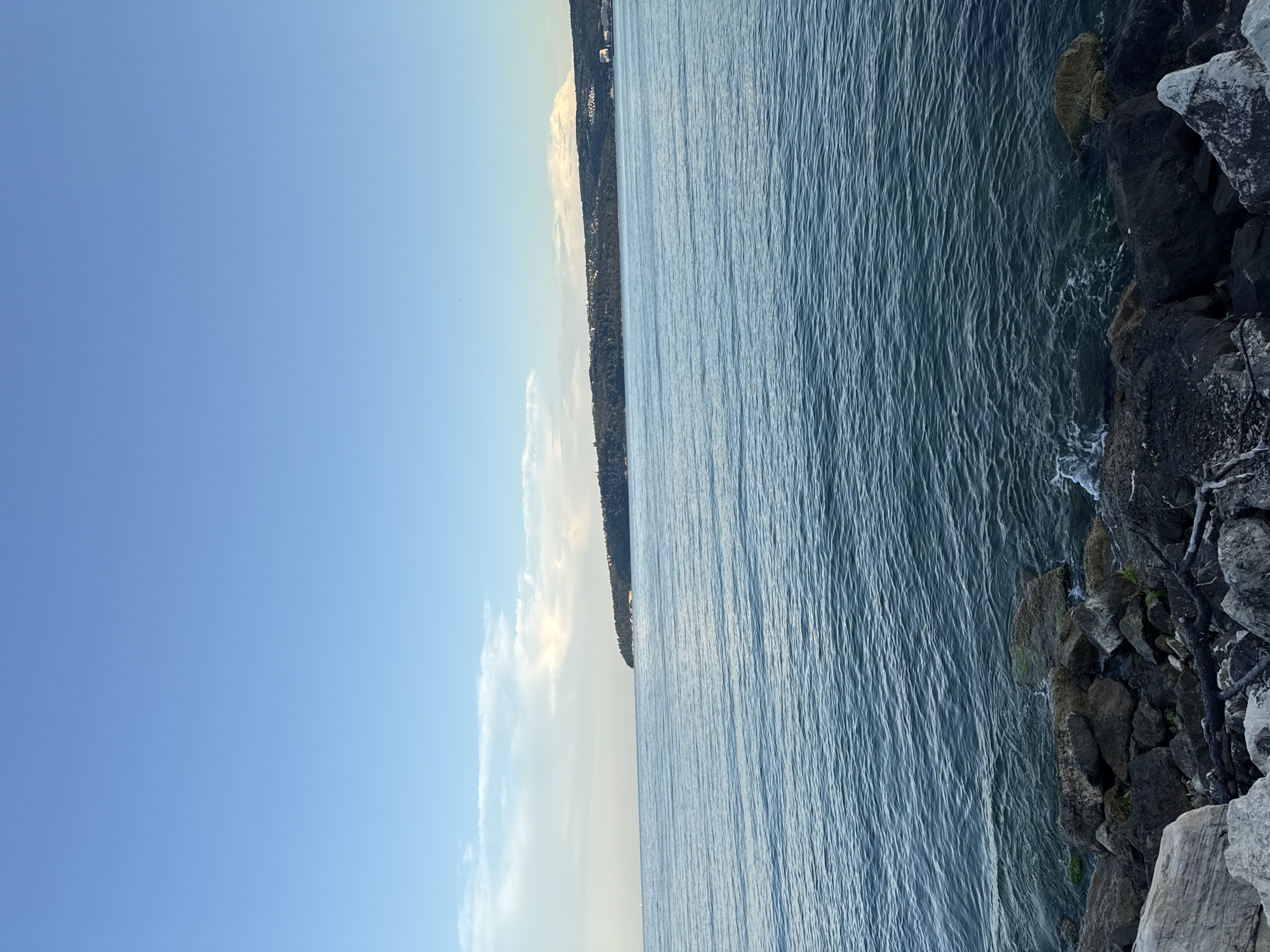 The image shows a body of water with a distant, forested coastline. The water is calm, with small ripples reflecting the light blue sky. The sky itself is mostly clear, with a few wispy white clouds on the horizon. Along the bottom of the frame, there is a rocky shoreline with dark, weathered stones. The scene suggests a natural landscape, likely a coast or a large lake, possibly during the late afternoon or early evening given the soft lighting. There are no people or animals visible in the frame.