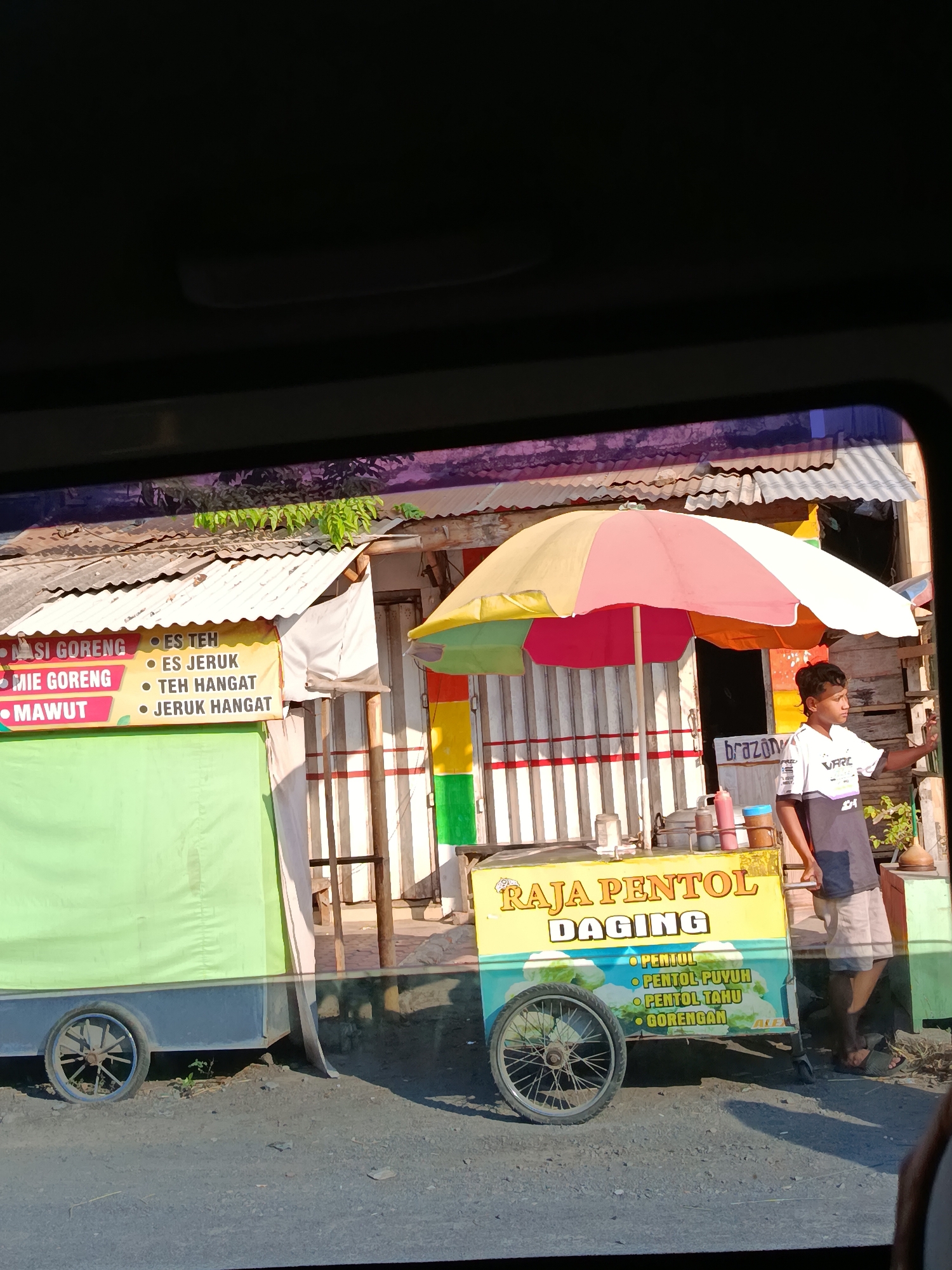 This image shows a street vendor in Wonosari, Indonesia, operating a food cart. The cart is adorned with colorful signage advertising "RAJA PENTOL DAGING" (King Meat Pentol), with a list of items including "PENTOL," "PENTOL PUYUH," "PENTOL TAHU," and "GORENGAN" (fried snacks). A young man, presumably the vendor, stands next to the cart, holding a smartphone and appearing to take a photo. The cart is equipped with an umbrella providing shade, and various containers and sauces are visible on its surface. Behind the cart, there is a building with a corrugated metal roof and brightly painted vertical stripes. The surrounding environment appears to be a roadside setting, with a dirt or gravel ground and a glimpse of greenery. The overall scene captures a moment of everyday commerce on a street in Wonosari.