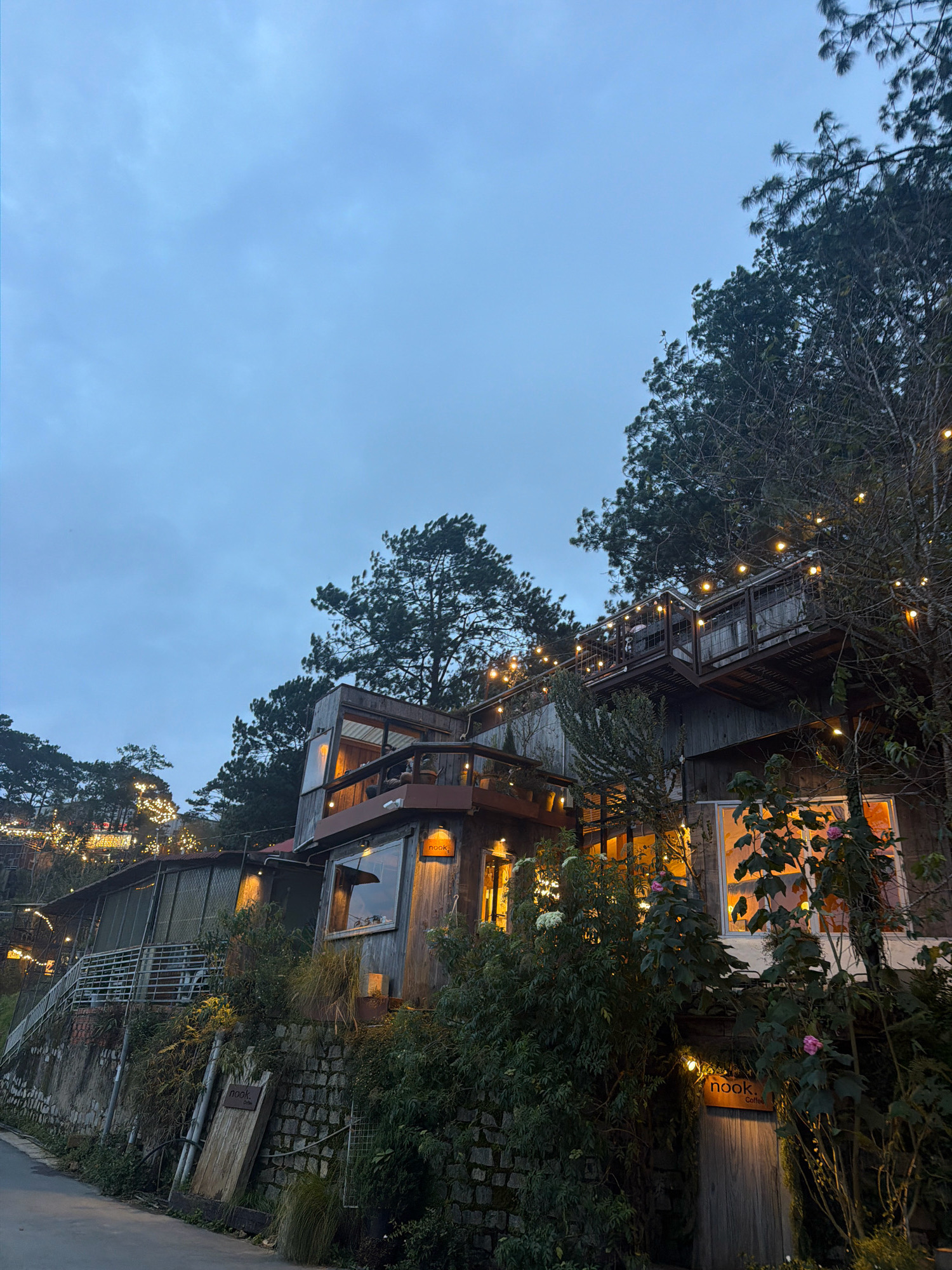 The image depicts a rustic, multi-level wooden building nestled on a hillside at dusk. The building is adorned with numerous warm-toned string lights, illuminating the exterior and highlighting the architectural details. The structure appears to be a cafe or guesthouse, with a sign near the entrance reading "nook. Coffee." Greenery, including trees and bushes, surrounds the building, with some plants reaching up towards the structure and lights. The sky is overcast and a deep blue, indicating either early evening or a cloudy day. A paved pathway or road is visible in the lower left quadrant of the image, suggesting it is accessible from this direction. The overall atmosphere is cozy and inviting, with the soft glow of the lights contrasting against the twilight sky and the natural setting. The location appears to be in the Xuân Trường - Đà Lạt area of Vietnam, known for its hilly terrain and pine forests, which are visible in the background.