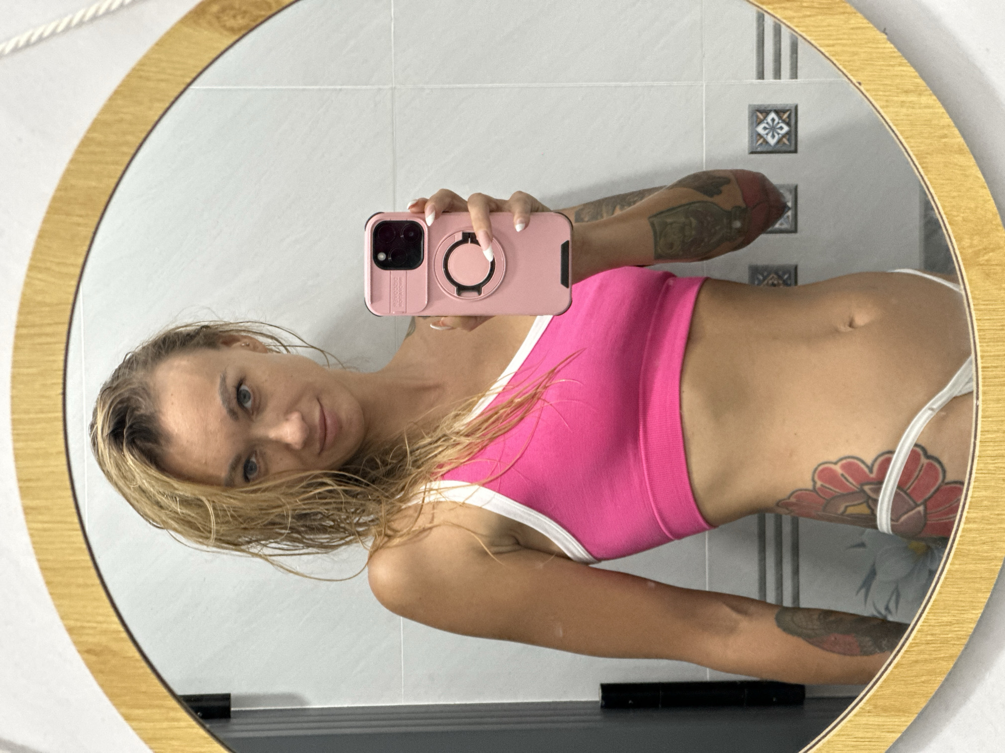 A woman with wet blonde hair, wearing a pink sports bra and white bikini bottoms, stands in front of a mirror in a bathroom. She is holding a pink phone in her right hand. Tattoos are visible on her left arm and hip. The bathroom has tiled walls and a wooden framed mirror. The scene is set in Mũi Né, Vietnam.