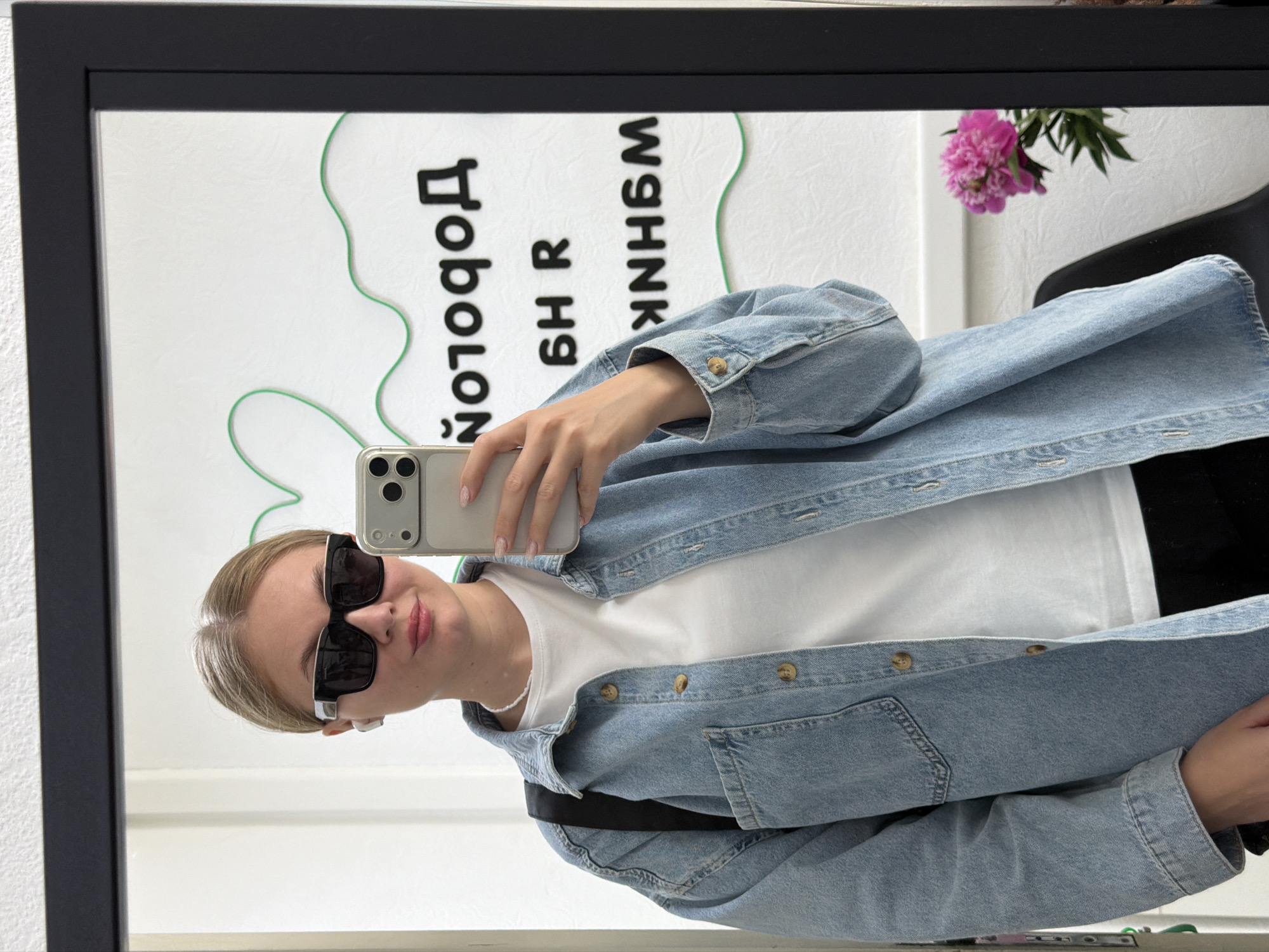 A young woman with blonde hair pulled back, wearing oversized black sunglasses, white earbuds, and a white t-shirt under a light blue denim jacket, takes a mirror selfie. She is holding a silver smartphone in her right hand. A black strap, possibly from a bag, is visible over her left shoulder. The background reflected in the mirror shows a white wall with abstract green designs and black text. A pink flower in a vase is visible to the right of the woman in the reflection. The scene appears to be indoors, possibly in a shop or studio. There is no specific indication of the city of Chaikovsky, Russia.