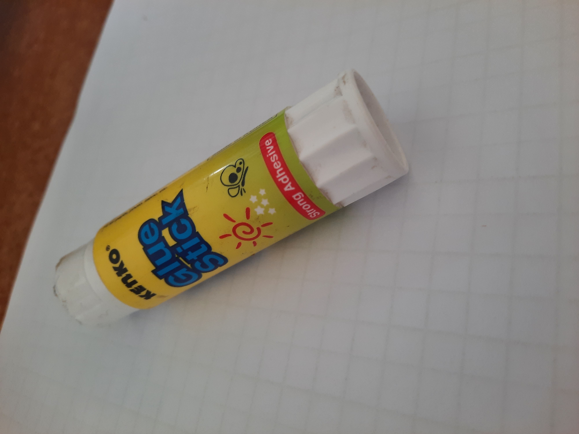 A close-up shot shows a yellow glue stick with blue lettering on a white grid paper. The glue stick has a white cap and a white base with ridges. The lettering on the glue stick reads "KENKO" and "Glue Stick" in a playful font. There is also a small illustration of a sun with rays and a butterfly. Below the illustration, the words "Strong Adhesive" are printed in a red oval shape. The glue stick is positioned diagonally across the frame, with the cap facing the top left and the base facing the bottom right. The background is a white paper with a faint grid pattern, suggesting it might be notebook paper. The lighting appears to be natural, casting soft shadows. The overall scene suggests a simple, everyday object placed on a desk or table, likely in a school or office environment. The image does not contain any people or specific landmarks that would indicate a location in Lhokseumawe, Indonesia.