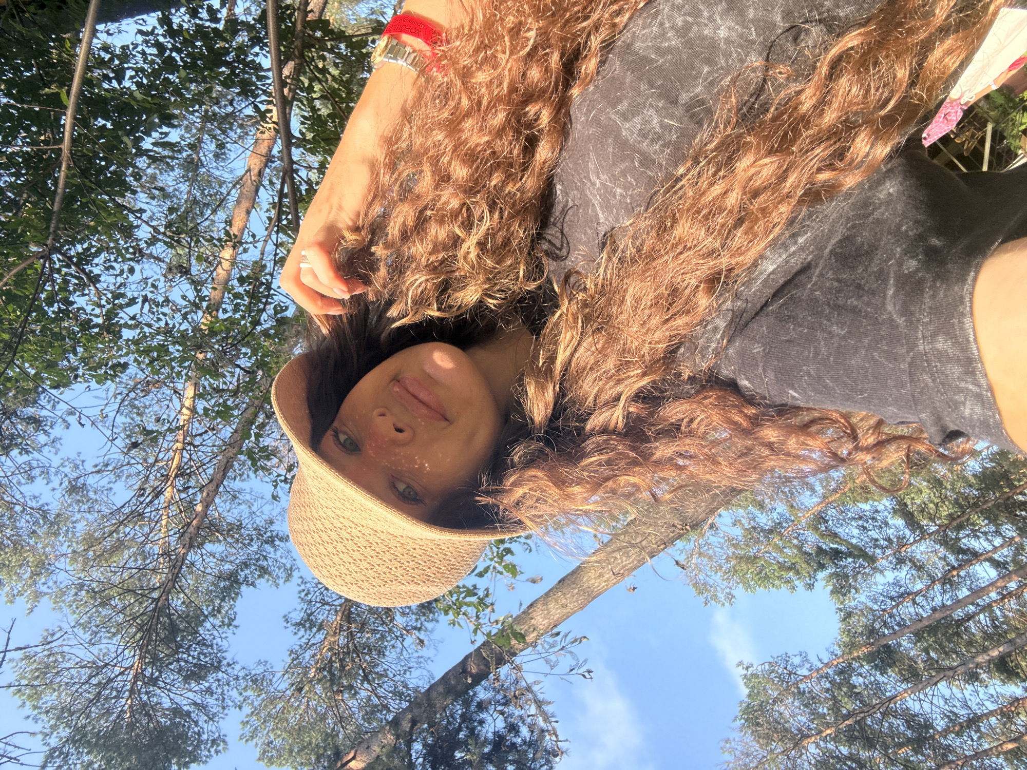 A young woman with long, curly auburn hair and a tan hat takes a selfie outdoors. She is wearing a dark grey t-shirt and a silver watch with a red band. The background consists of tall trees and a clear blue sky, suggesting a natural setting. The lighting indicates it is daytime. The location is identified as Barangol, Russia.