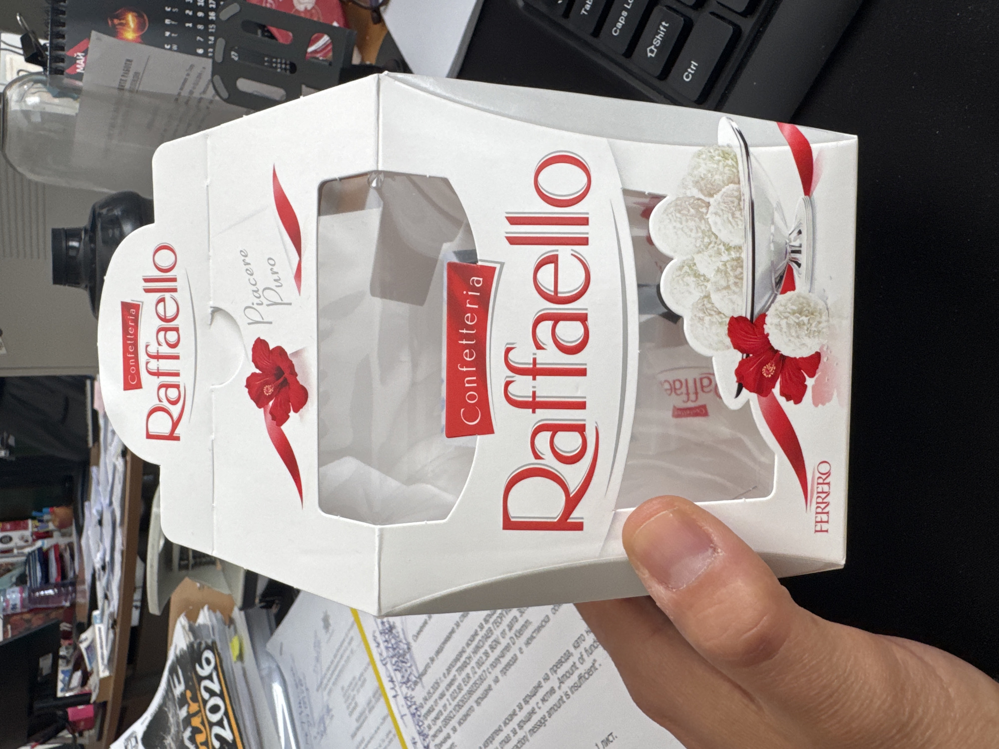 A hand holds a box of Raffaello candies. The box is white with red lettering and images of the candy inside. The background includes a keyboard, a calendar, and papers with text in Bulgarian. The text refers to refunds and insufficient funds. The year 2026 is visible on one of the documents. The location seems to be an office in Plovdiv, Bulgaria.