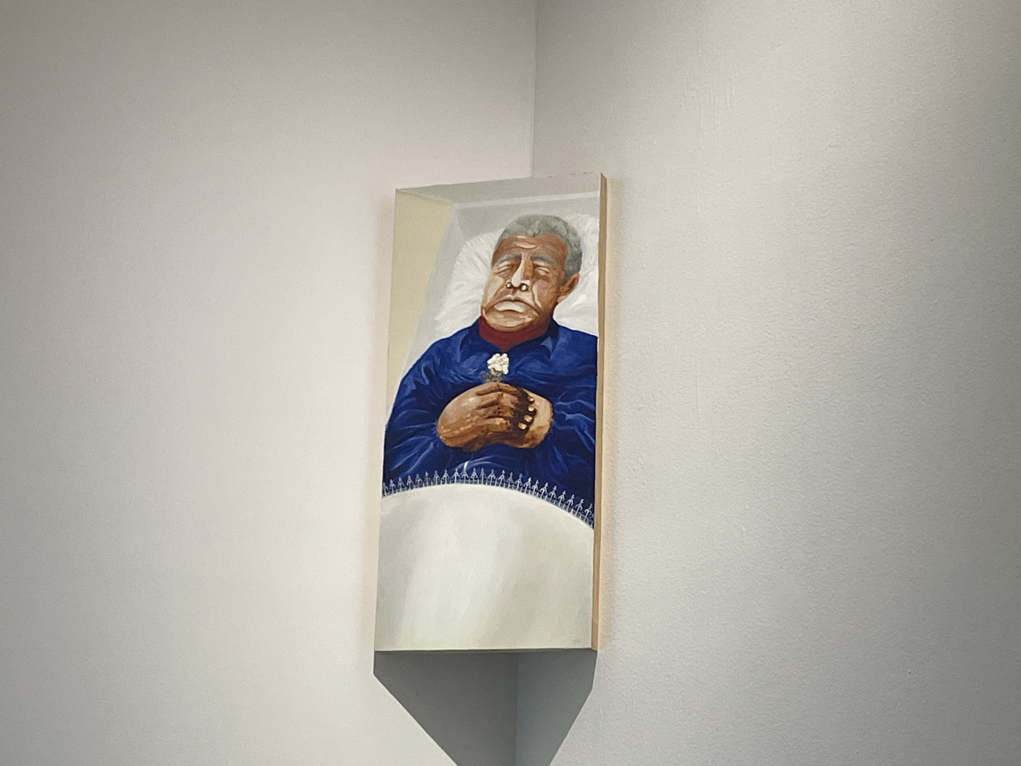 A rectangular painting hangs on a light gray wall in the corner of a room. The painting depicts an older man, likely deceased, lying in what appears to be a coffin or bed. He has gray hair and is wearing a dark blue shirt. His hands are folded together, holding a small white flower. The man lies on a white sheet with a decorative border. The location is inferred to be Mexico City, Mexico, based on image content and source data.