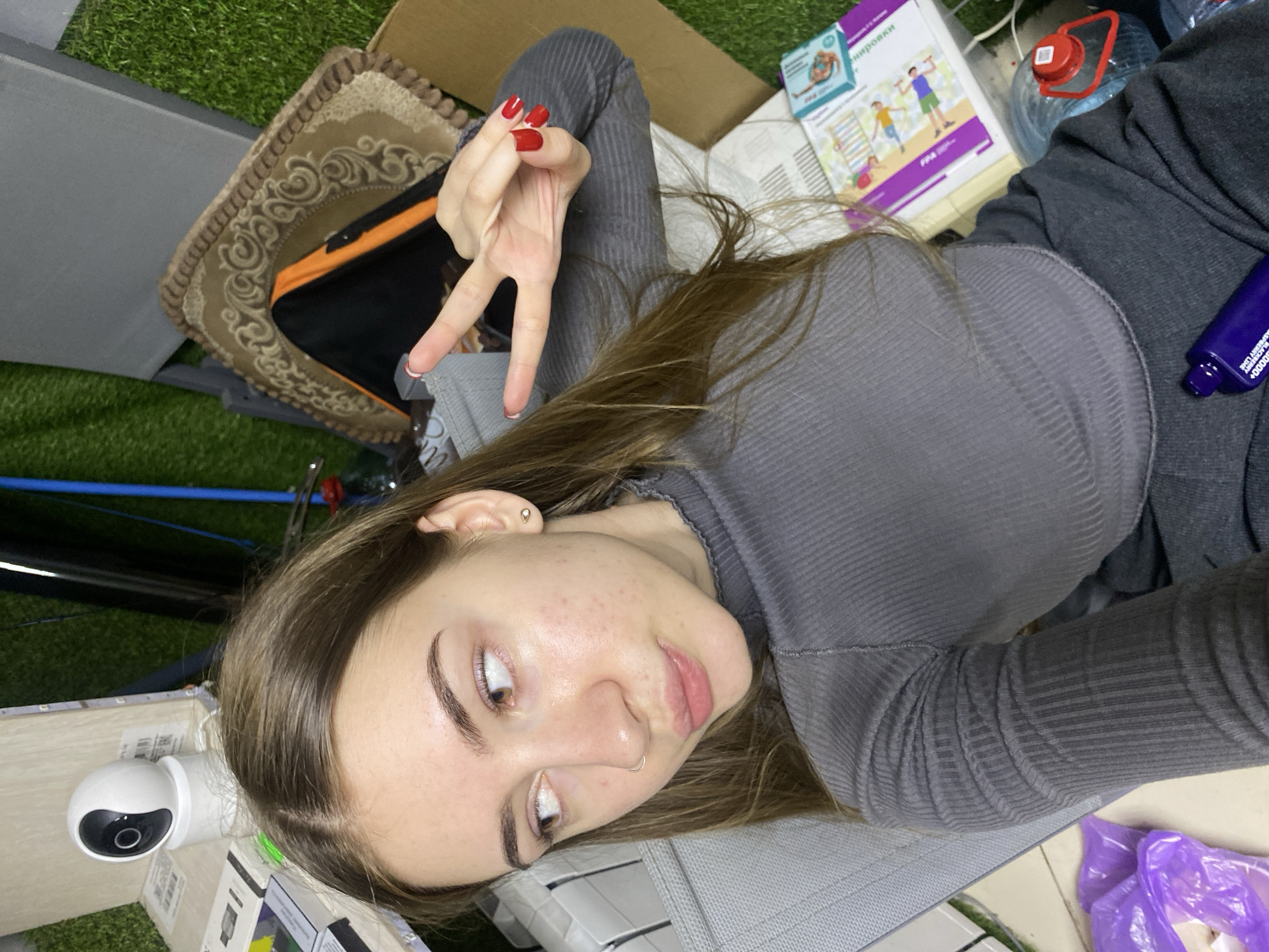 A young woman with long brown hair is pictured from the chest up, facing right and looking away from the camera. She is wearing a grey, ribbed, long-sleeved top. Her right hand is raised, forming a peace sign, with red nail polish visible on her fingernails. The background appears to be indoors, with a grassy artificial turf visible behind her and various objects scattered around, including what looks like a white security camera, boxes, and a roll of dark material. A book with colorful illustrations is also present on a surface to her right. The overall impression is of a casual, informal setting. There is no visible indication of the location being in the city of Дьяково, Russia.