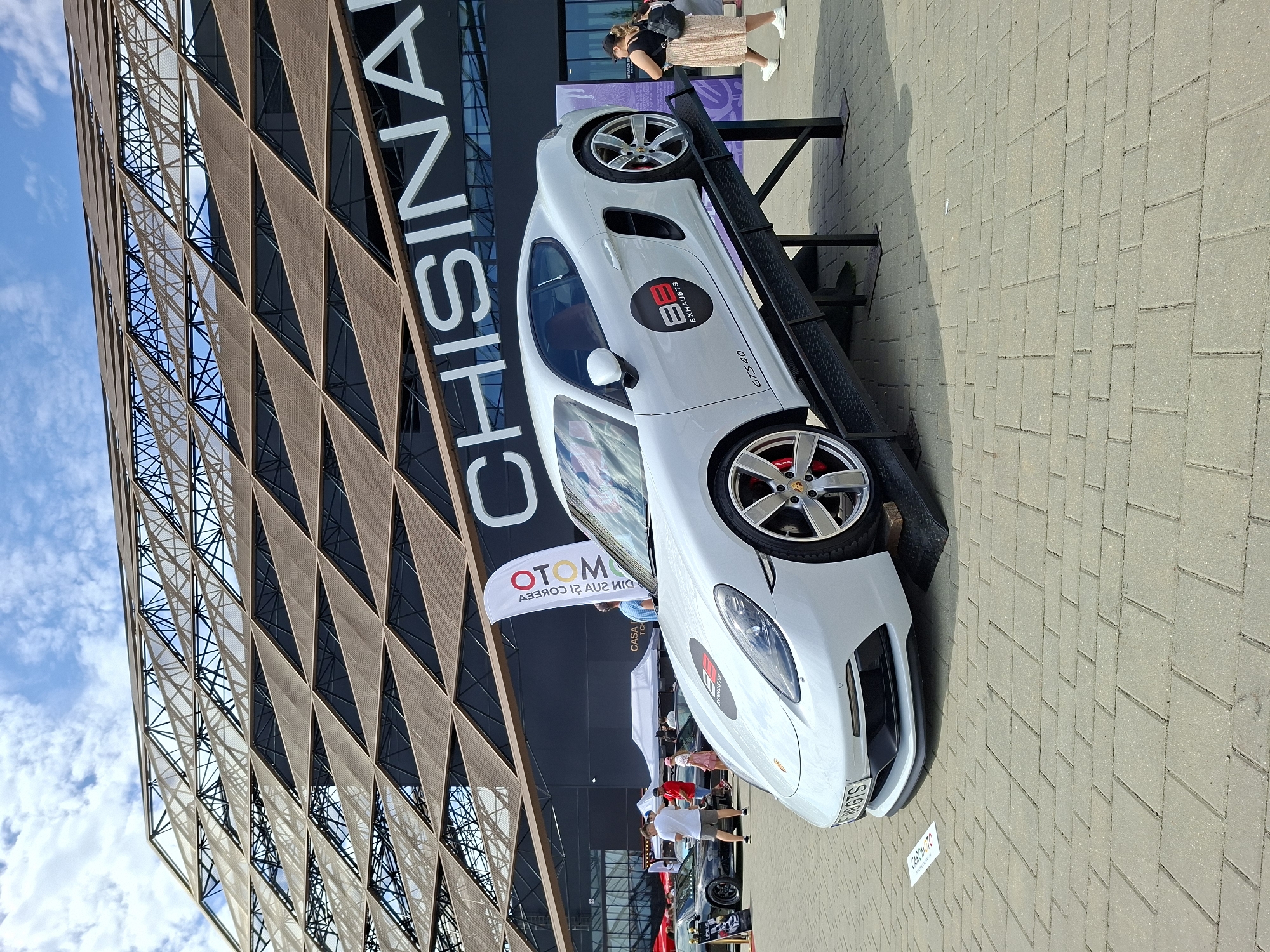 A white Porsche GTS 4.0 is displayed on a ramp outside the Chisinau Arena in Stăuceni, Moldova. The car has a red brake caliper and a black circular logo with "BB EXHAUSTS" on the driver's side door. A license plate reads "98 GBGTS". The background features the distinctive patterned facade of the Chisinau Arena and a blue sky with scattered clouds. Several people are visible in the background, some walking and others standing near tents and other vehicles. A white flag with "OTOM" written on it stands near the car. The ground is paved with gray interlocking bricks. The overall scene suggests an outdoor event or car show.