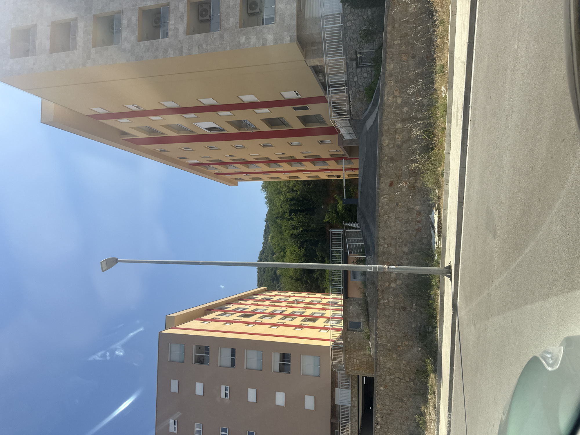 The image shows two apartment buildings on a sunny day in Boreti, Montenegro. The building on the left is light brown with a yellow top and red accents on its facade. It has numerous windows, some with white shutters. The building on the right is predominantly yellow with red vertical stripes and a grey stone-clad section. It also features multiple windows and balconies. Both buildings appear to be multi-story residential structures.
A tall, grey lamppost stands in the foreground, positioned between the two buildings. The buildings are situated on a slope, with a retaining wall made of rough stones in front of them. There is a paved area and a sidewalk in front of the wall, with a road visible in the lower part of the image, suggesting the perspective is from a vehicle. Lush green trees and vegetation cover the hillside behind the buildings, indicating a somewhat rural or semi-urban setting. The sky is clear blue with some faint white streaks, possibly contrails. The overall scene depicts a residential area on a clear, bright day, likely in a hilly or mountainous region given the background.