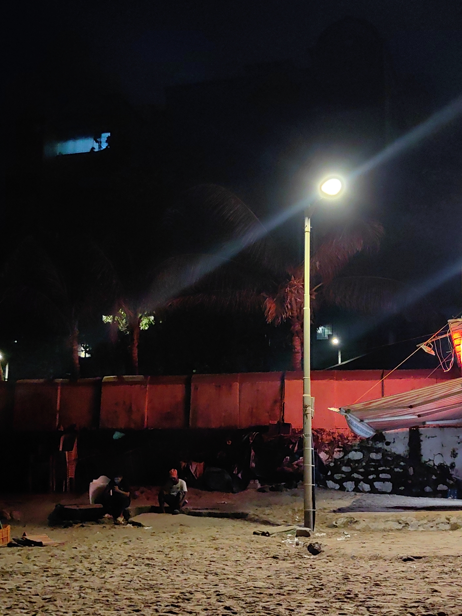 The image depicts a nighttime scene on a sandy beach in Mumbai, India, illuminated by a street lamp. In the foreground, the sandy ground is visible, with some debris scattered around. A prominent street lamp stands in the center of the frame, casting a bright light. In the mid-ground, a low concrete wall runs horizontally, painted a reddish-orange hue. Behind the wall, palm trees are silhouetted against the dark sky. Several people are visible in the mid-ground, some appearing to be sitting or squatting. One person in the lower right section of the image is wearing an orange cap. To the right of the street lamp, a striped canopy or shelter is partially visible, supported by a rope. In the background, the sky is dark, with some faint lights suggesting urban activity in the distance. The overall atmosphere is dimly lit and appears to be a public outdoor space at night.