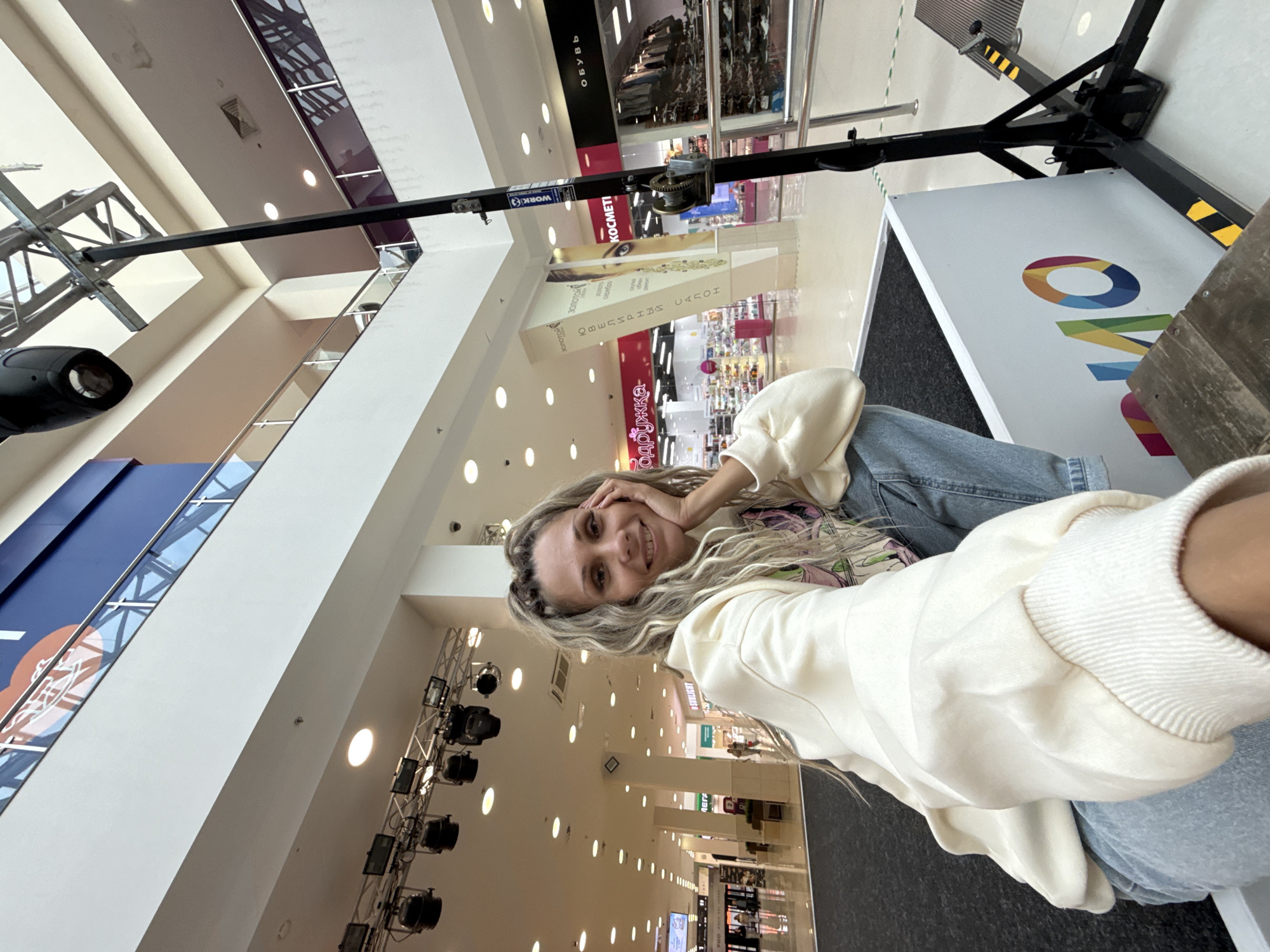 A woman with blonde braided hair smiles at the camera, resting her chin on her hand. She is wearing a cream-colored hoodie and blue jeans. She is seated on a raised platform in what appears to be a shopping mall. The background shows a multi-level mall interior with shops, escalators, and lighting. A black lighting rig with multiple spotlights is visible above and to the left of the woman. A large white structure with colorful lettering is in the foreground to her right.