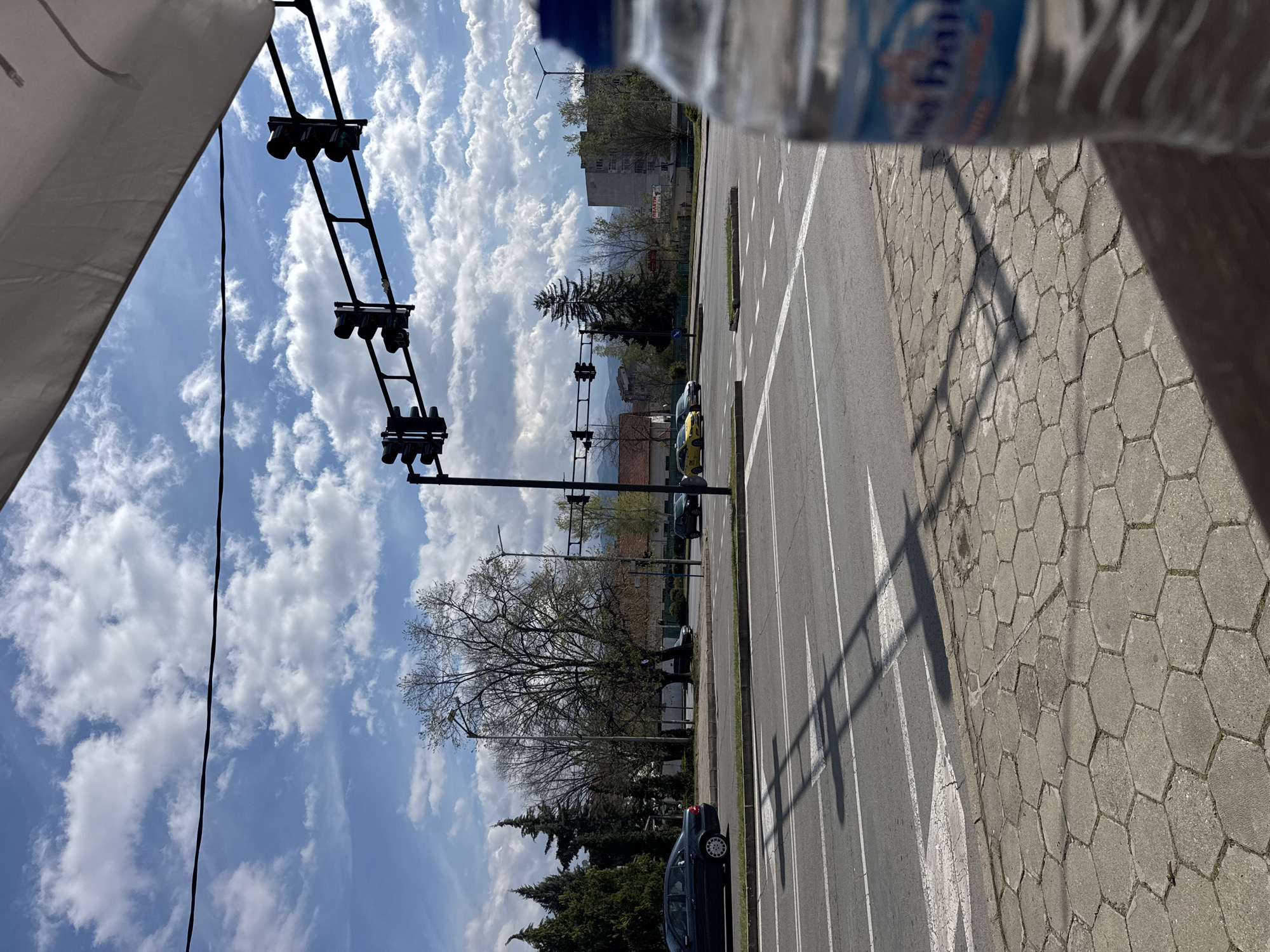 A street scene in Blagoevgrad, Bulgaria, shows a multi-lane road with a crosswalk and multiple traffic lights. Cars are parked along the side of the road. A sidewalk with hexagonal paving stones is adjacent to the road. A bottle partially obscures the view at the top of the image. The sky is blue with scattered white clouds. Buildings and trees are visible in the background.