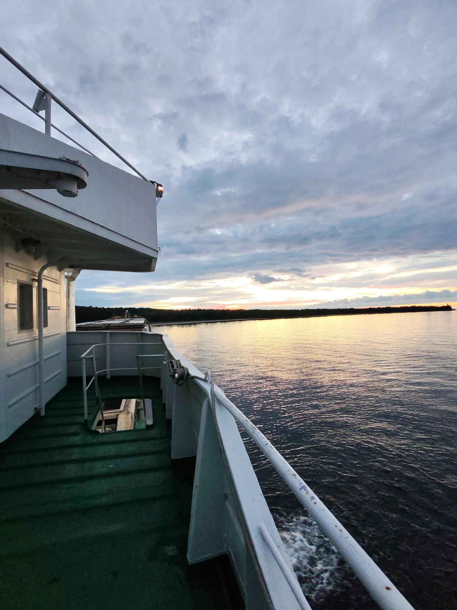The image captures a view from the deck of a ferry boat traveling on a body of water, likely a river or lake, near the city of Shcheleiki, Russia. The perspective is from the side of the vessel, looking forward and to the right. The sky is overcast with dramatic clouds, tinged with the soft hues of either sunrise or sunset, casting a warm glow on the water's surface. The water is dark and rippled, with a white wake trailing from the ferry, indicating movement.
Dominating the left side of the frame is the white superstructure of the ferry, including a railing, windows, and a portion of the deck covered in green, textured material. The deck shows some wear and tear, with a visible hatch or opening leading to a lower level. On the right, the railing of the ferry extends into the frame, showing signs of rust and peeling paint.
In the distance, a dark, tree-lined shore stretches across the horizon. The low light suggests it is either early morning or late evening. There are no people visible in the image. The overall atmosphere is calm and somewhat solitary, conveying a sense of travel and the vastness of the natural environment surrounding the ferry.