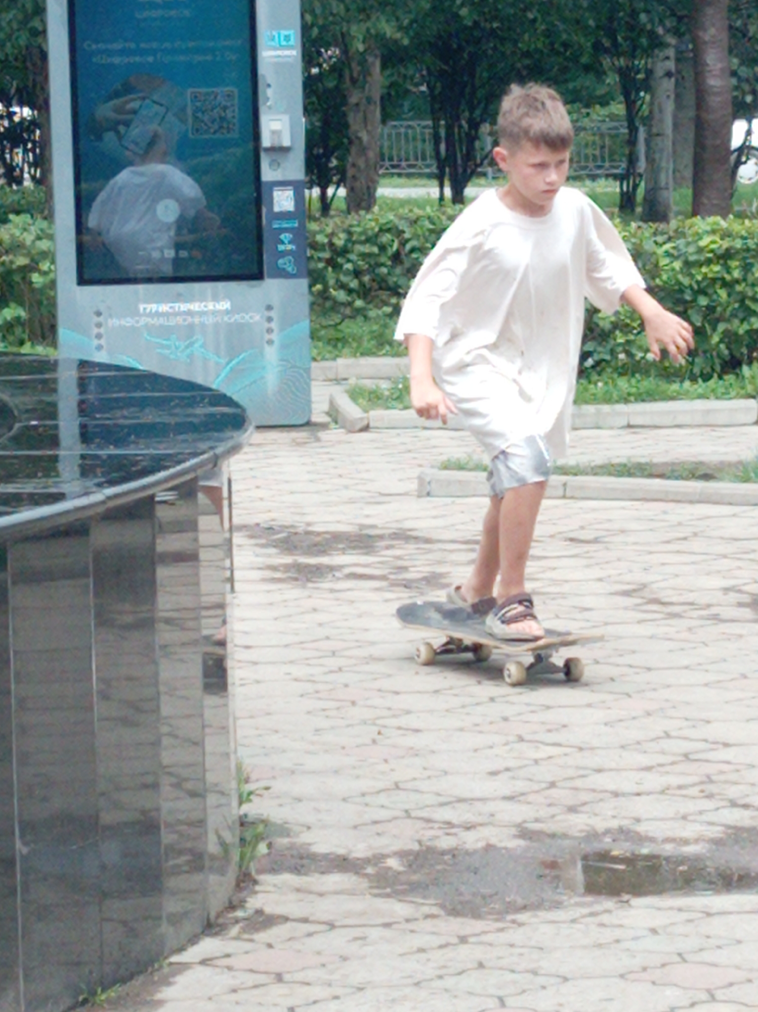 A young boy is skateboarding on a paved pathway in a city park in Artyom, Russia. The boy, appearing to be around 10-12 years old, is wearing a loose white t-shirt and silver shorts, with sandals on his feet. He is actively engaged in skateboarding, with his body angled forward and his hands extended for balance. The skateboard is a standard model with visible wheels and trucks.
The environment suggests a public outdoor space. Behind the boy, a large digital information kiosk stands, displaying graphics and text in Russian. This kiosk is typical of those found in tourist areas or public squares. To the left of the scene, a dark, reflective structure, possibly a bench or a planter, occupies a significant portion of the frame. The pathway is made of interlocking pavers and shows signs of recent rain, with several puddles present. In the background, trees and green foliage indicate a park-like setting. The overall impression is a snapshot of a casual summer day activity in a Russian city.