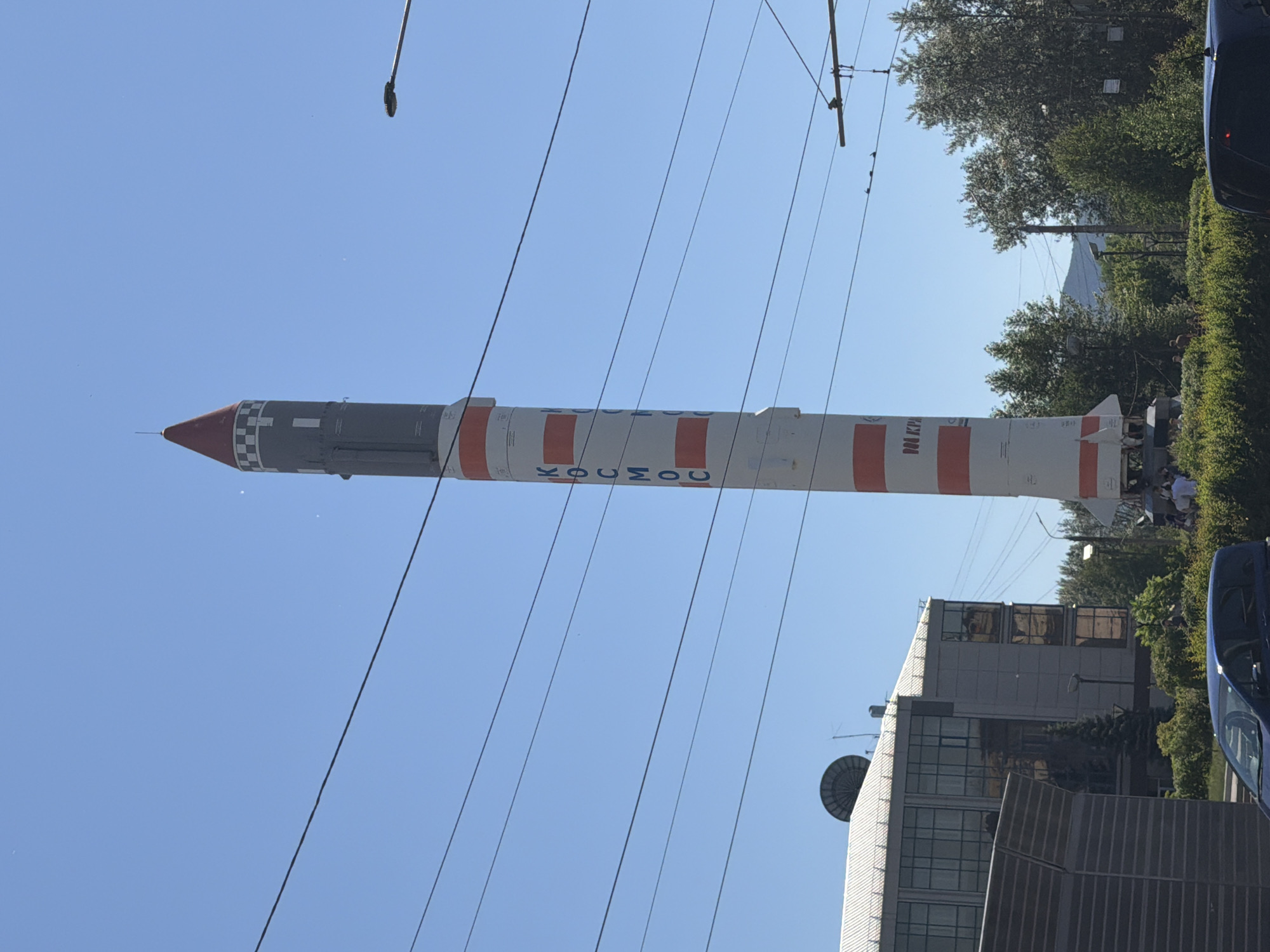 The image shows a monument to a rocket in the city of Krasnoyarsk, Russia. The rocket is painted white and red, with the word "КОСМОС" (KOSMOS) written vertically in blue letters on its side. It is a tall, slender structure, tapering to a pointed red and white tip at the top. Below the rocket, a group of people are gathered. In the background, there is a building with a large satellite dish on its roof, as well as trees and a clear blue sky. Overhead power lines are also visible, suggesting the presence of public transportation like trams or trolleybuses. The scene appears to be a public space, likely a park or a square, given the monument and the gathering of people.