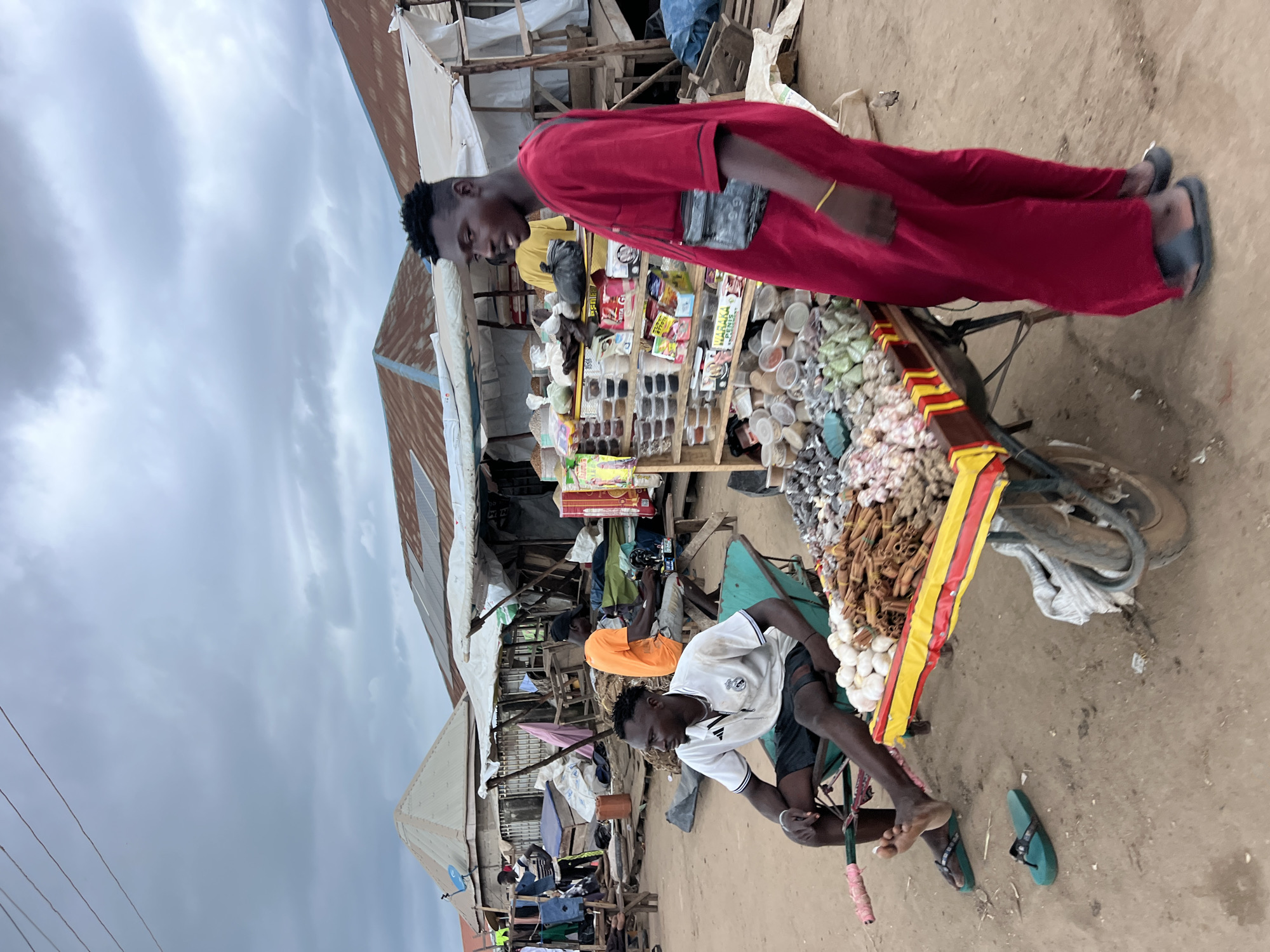 In Oko Amakom, Nigeria, under a cloudy sky, an outdoor market scene unfolds. A man in a red robe stands near a cart laden with produce, including garlic and cinnamon sticks. To his left, a man in a white polo shirt and shorts sits in a green wheelbarrow, wearing sandals. Another man in an orange t-shirt operates a sewing machine at a wooden stall in the background. The market consists of makeshift stalls with corrugated tin roofs, some with tarpaulins. Various goods, including packaged items and sacks of produce, are displayed. The ground is composed of dirt and sand.