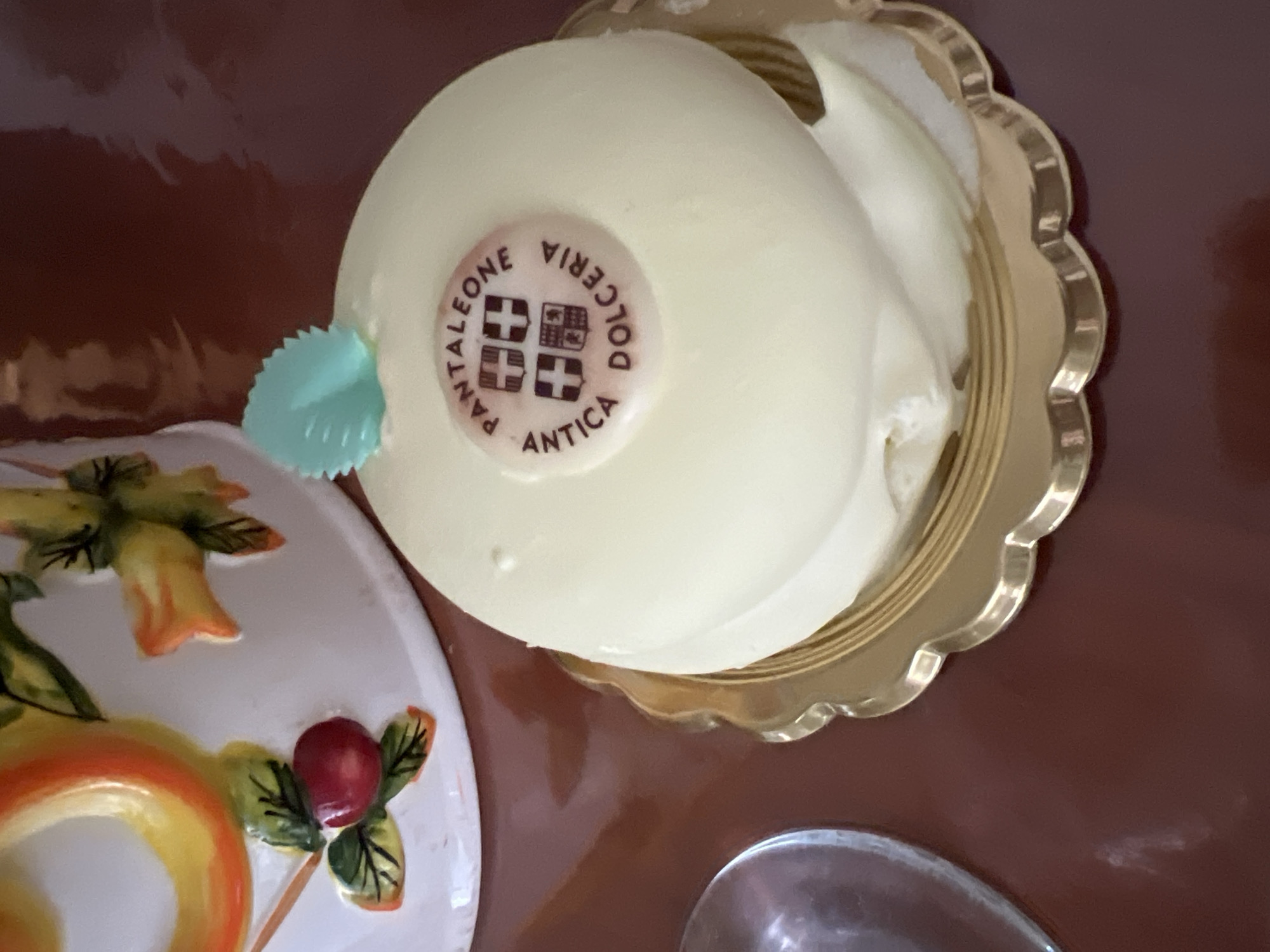 The image shows a small, round, white cake with a light green decorative leaf on top, placed on a golden fluted plate. A circular logo is visible on the cake, featuring text that reads "PANTALEONE ANTICA DOLCERIA" and a crest with four quadrants. The cake is positioned on a reflective, dark brown surface, possibly a table. In the background, to the left, is a portion of a white ceramic object decorated with colorful fruit motifs, specifically an orange spiral and a red berry with green leaves. The overall impression is that of a dessert displayed for sale or consumption. The "Antica Dolceria Pantaleone" is a well-known pastry shop located in Salerno, Italy.