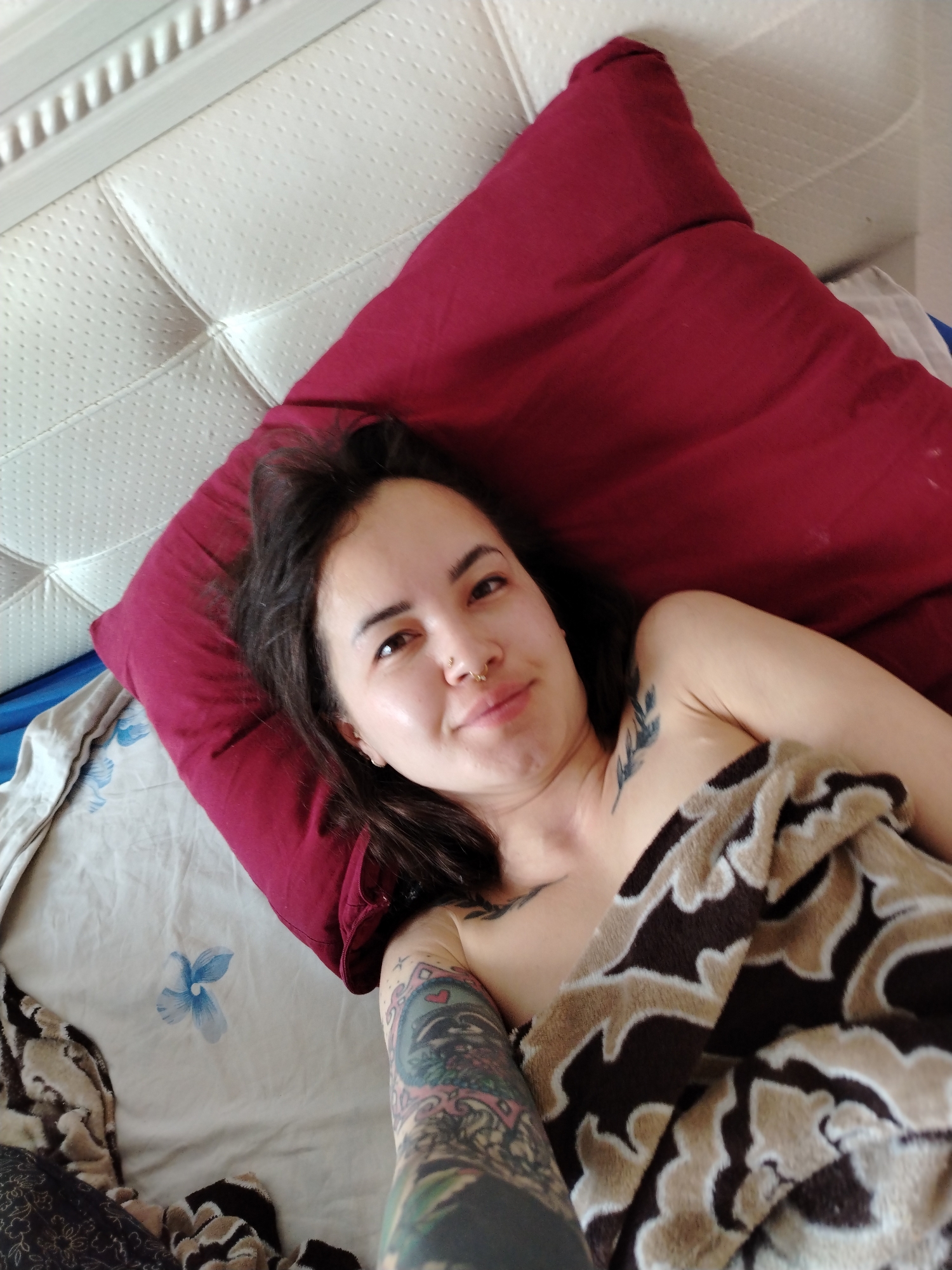 A woman with dark hair and a nose piercing lies in a bed with a red pillow behind her head. She is partially covered by a patterned blanket in shades of brown and beige. Her left arm, which is heavily tattooed, is visible. The bed has a white headboard with a textured pattern. The bedsheets have a faint floral pattern. The location in the city of Noviy, Russia is unknown.