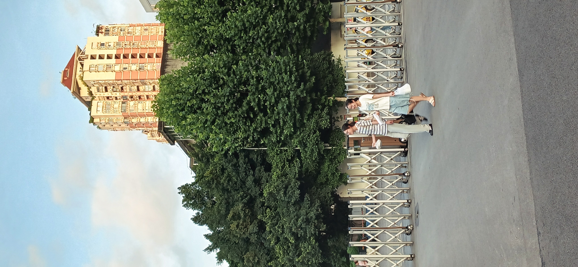 Two individuals walk along a grey asphalt surface bordered by a collapsible metal fence, near lush green trees and a tall building with multiple windows. The sky is light blue. The setting appears to be a street scene, likely in an urban area. The building's architecture suggests a location in Yuzhong District, China.