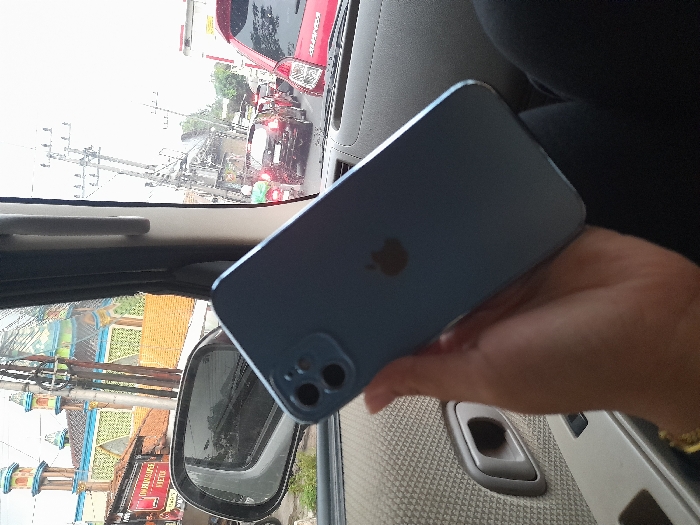 The image depicts an interior view from a vehicle in Tunggorono, Indonesia, looking outwards through the front passenger window and windshield. In the foreground, a left human hand, wearing a gold bracelet on the wrist, holds a light blue Apple iPhone, showcasing its rear side with the prominent Apple logo and dual-camera system. The hand is positioned against a beige or light grey patterned fabric car door panel, which features a grey door handle and power window switch.
Through the vehicle's windows, an urban street scene is visible under an overcast sky. Traffic appears congested, with multiple vehicles stationary or moving slowly in a line. A red Mitsubishi Xpander is positioned directly ahead of the viewer's vehicle, and other cars, including a dark SUV, along with motorcycles, are present further down the road. Utility poles with numerous wires line the street.
To the left, beyond the vehicle's side-view mirror, a mosque with two distinctive green and gold minarets and a geometrically patterned green and blue roof is clearly visible. Adjacent to the mosque stands a smaller building with an orange tiled roof, featuring a partially obscured red sign that reads "MAKANAN."
